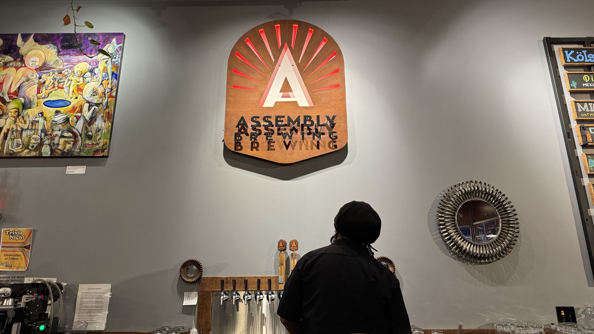 A man pours a beer from a tap against a wall with a large Assembly Brewing sign overhead.