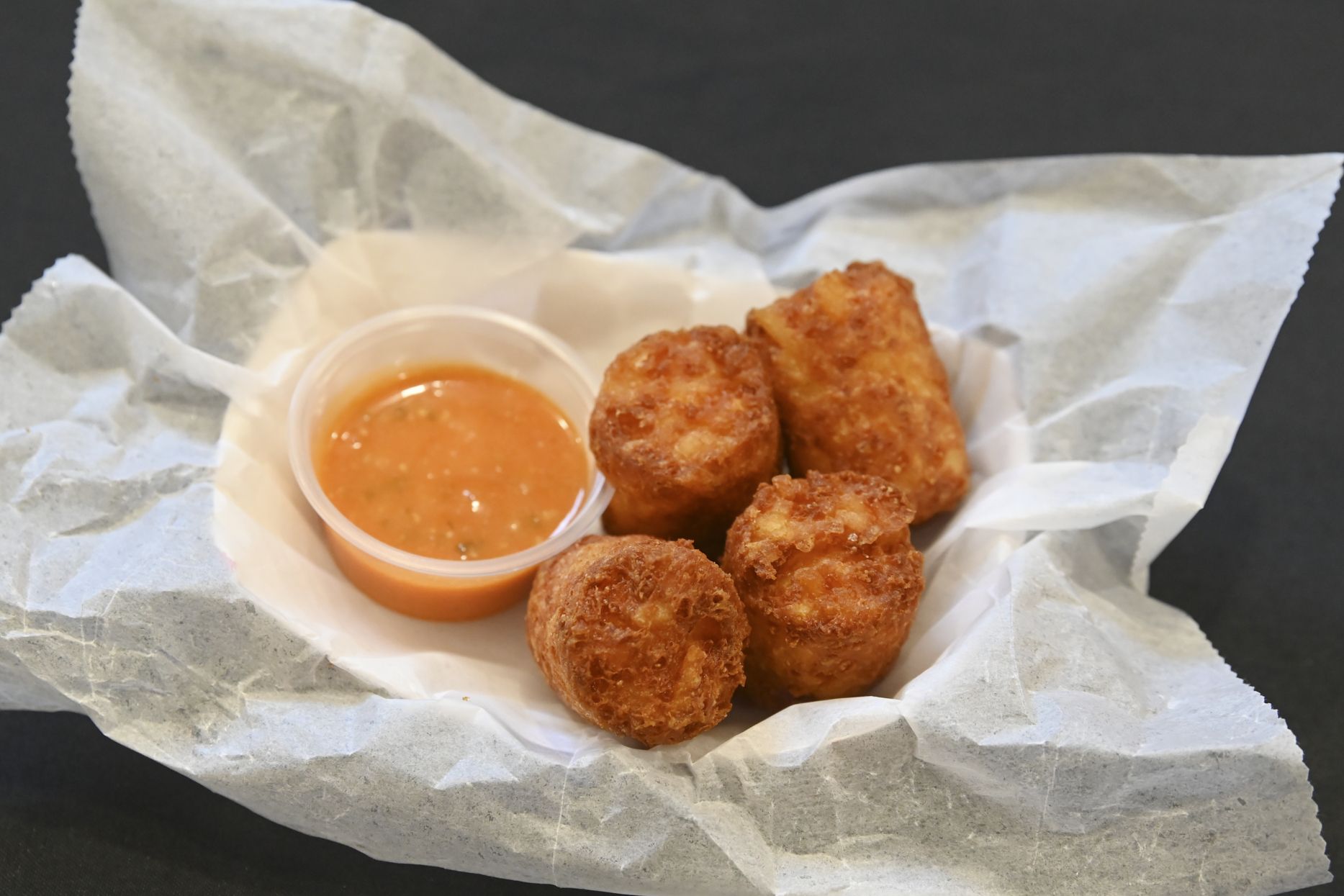 Golden-brown, breaded fried bites with a small cup of orange dipping sauce, set on crumpled parchment paper against a dark background.