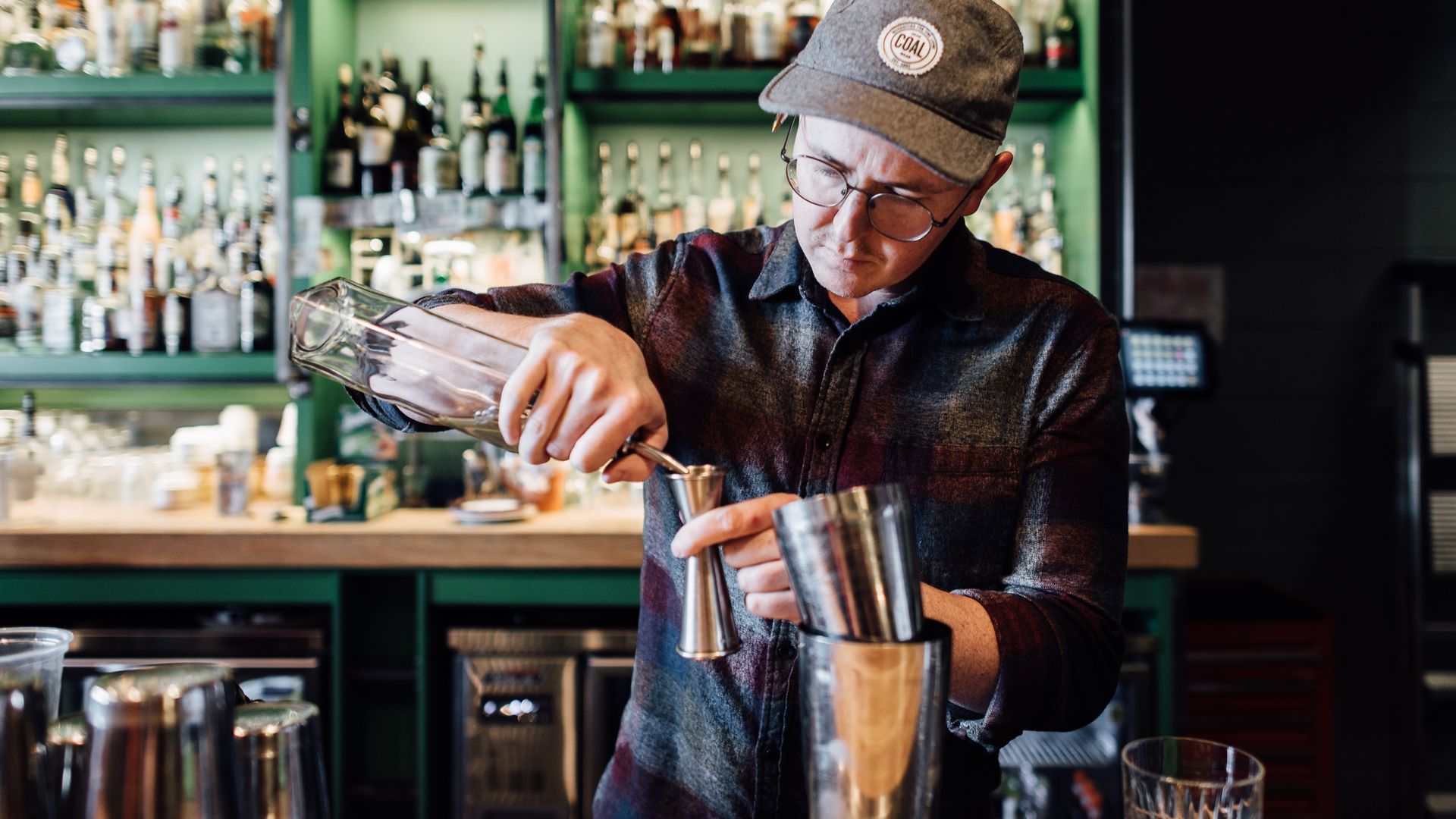 A bartender in a baseball cap pours a drink into a jigger.