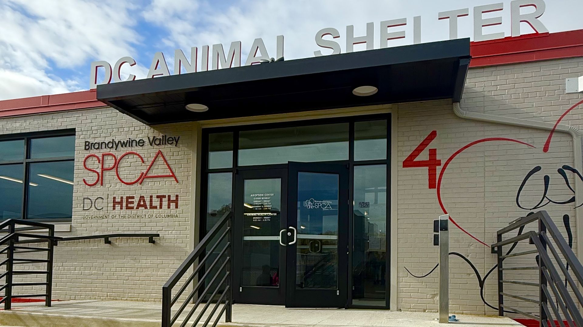 Entrance of the DC Animal Shelter with brick walls, black double doors, stairs, and signs for Brandywine Valley SPCA and DC Health under a partly cloudy sky.