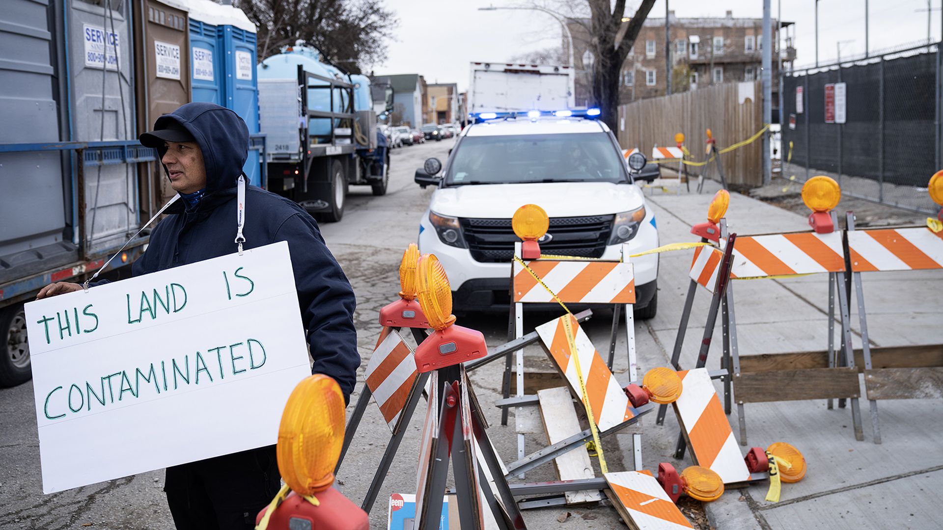 Man holding sign reading "This land is contaminated" in front of a police SUV and next to traffic blockades. 