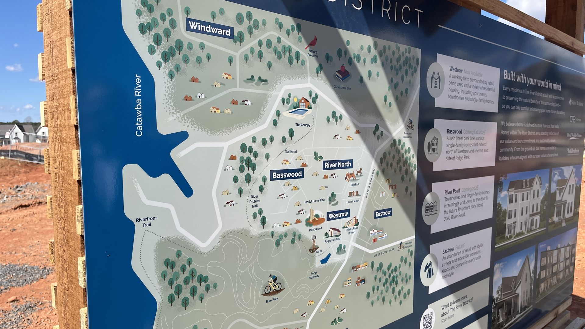 Bright blue outdoor sign with illustrated map of a nature district. Shows river on left, forest, houses, winding trails, labeled areas like Basswood and River North, plus right-side info panels.
