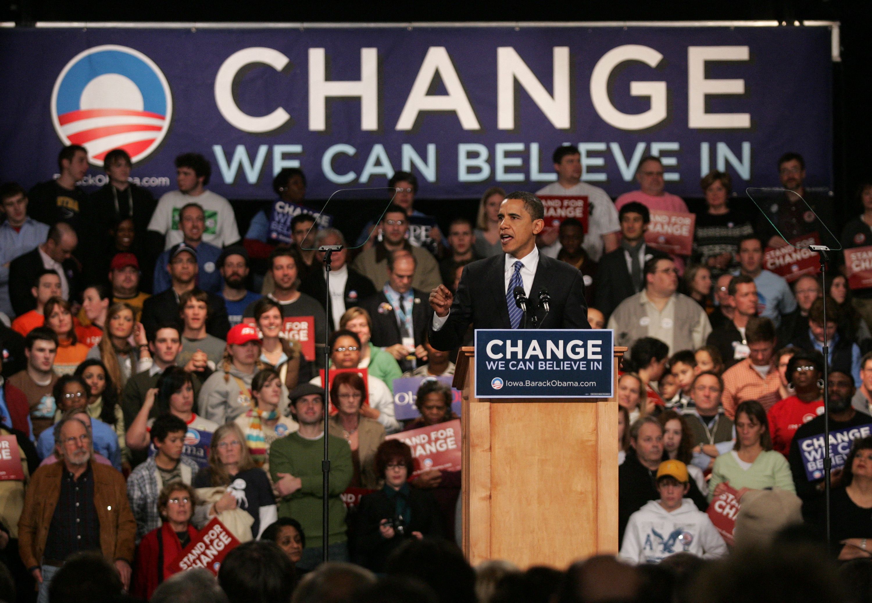 Obama stands at a lectern with a crowd behind him as he gives a speech. A sign says "CHANGE we can believe in" 