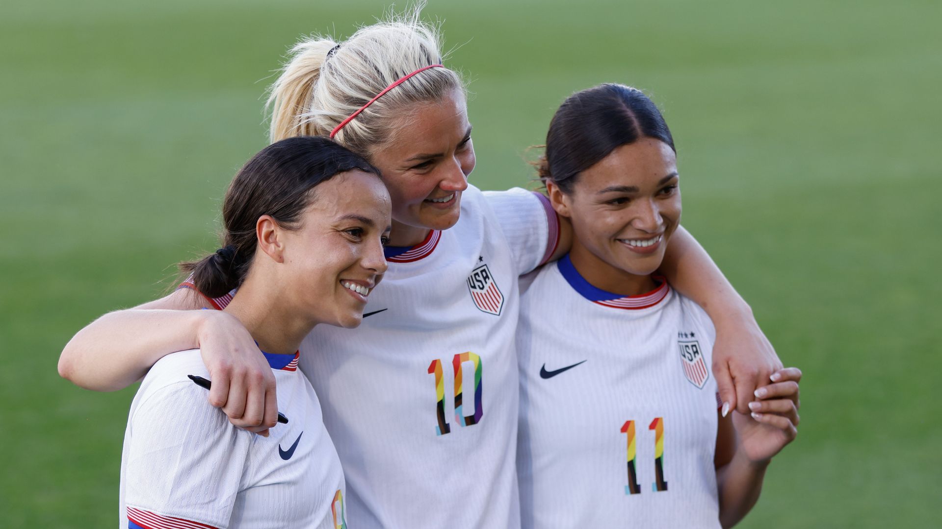 Three women in white jerseys smile while posing for a photograph while standing on a field. 