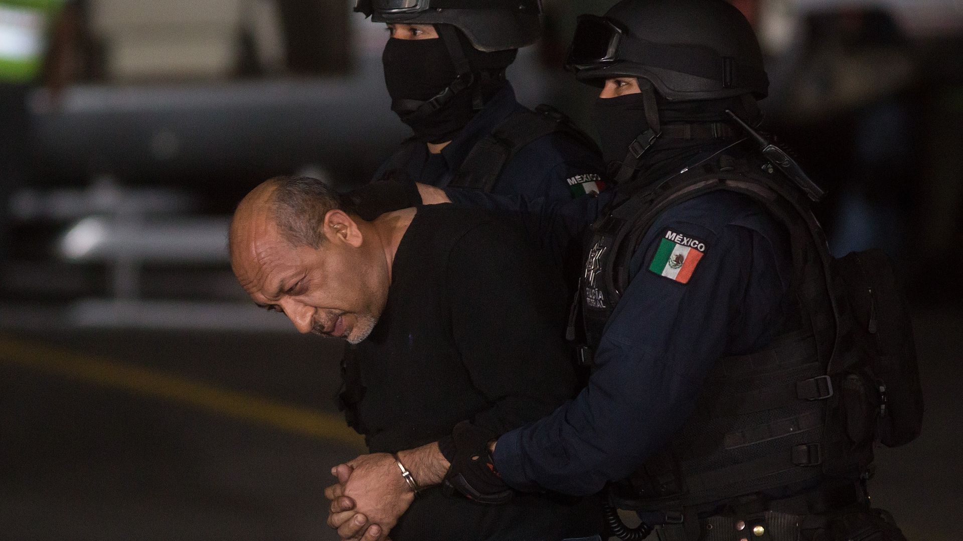 Two armed Mexican police officers in black helmets and masks arrest a handcuffed man in a black shirt, holding him from both sides in a dimly lit setting.