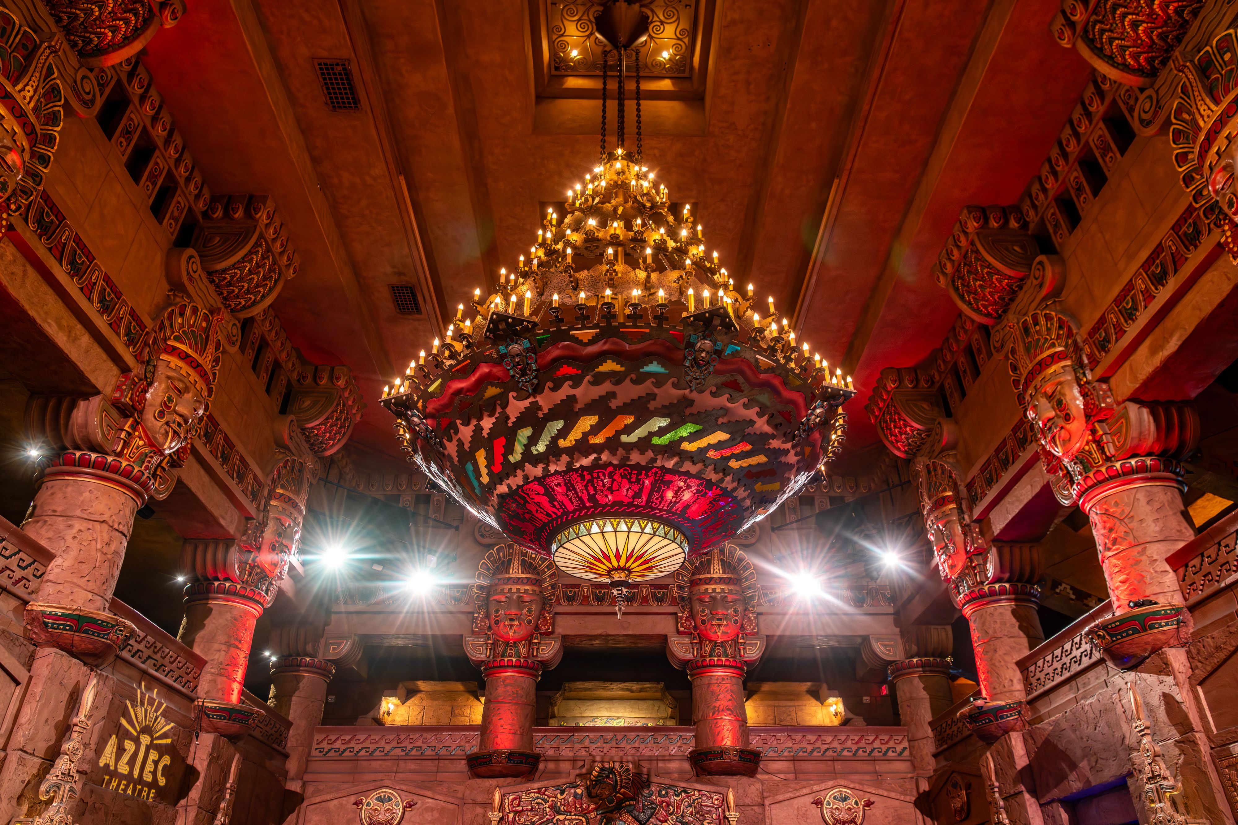 Inside an ornate theater, a grand multi-tiered chandelier lights a warm red-gold hall. Aztec-inspired columns and carvings line the walls, with a visible sign reading AZTEC THEATRE.