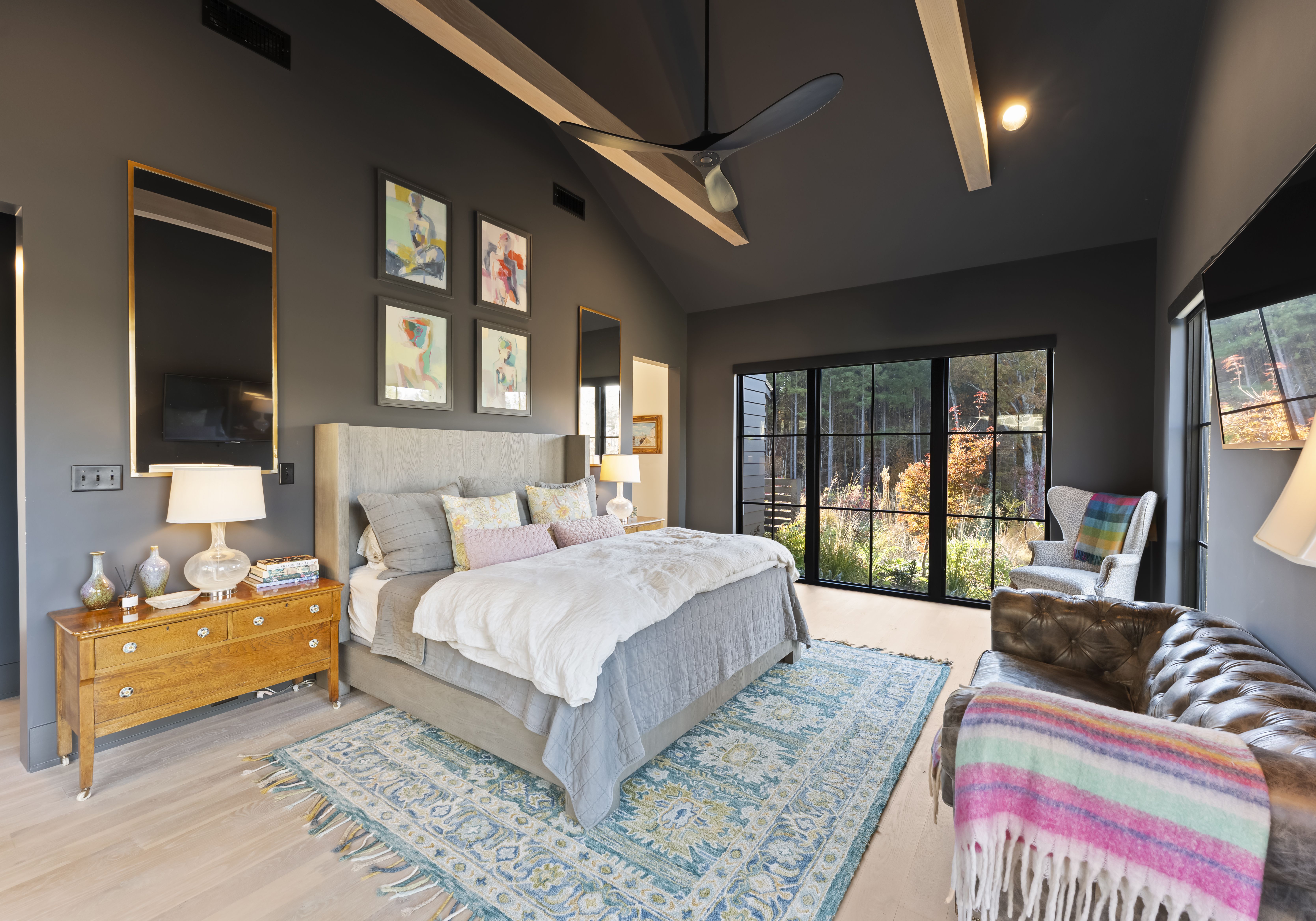 Modern bedroom with dark gray walls, a large gray upholstered bed in white bedding with pastel pillows. A blue patterned rug, brown leather sofa, a ceiling fan, and sliding glass doors to a sunny garden.