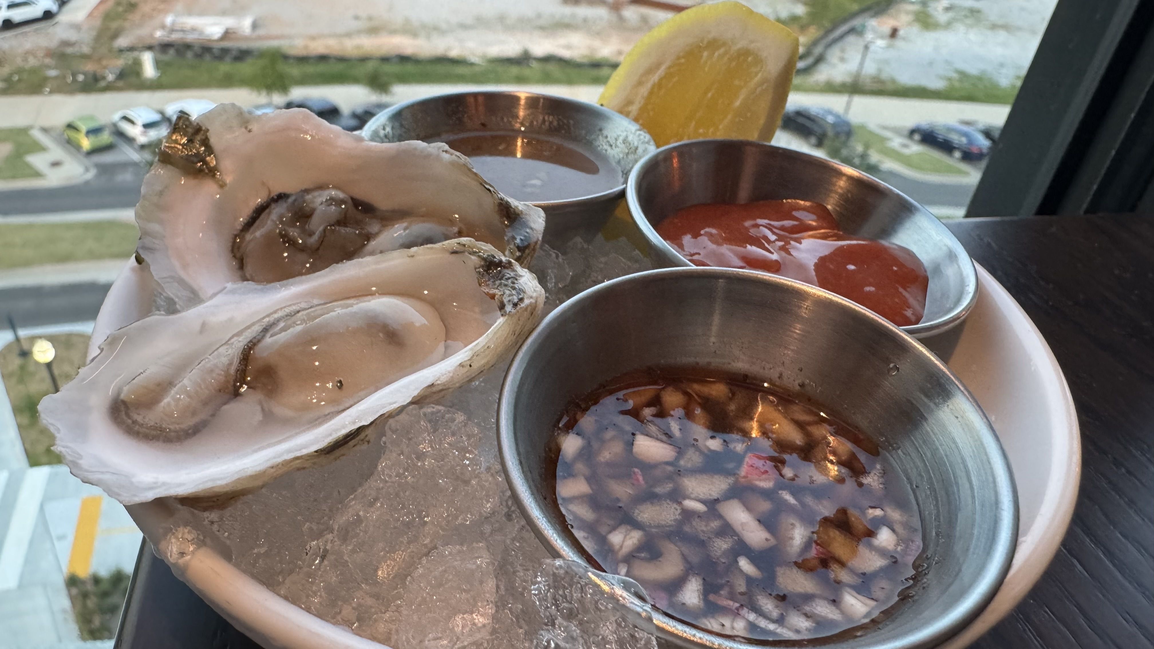 Two raw oysters on the half shell are served on ice with cocktail sauce, mignonette, a lemon wedge, and another dipping sauce.
