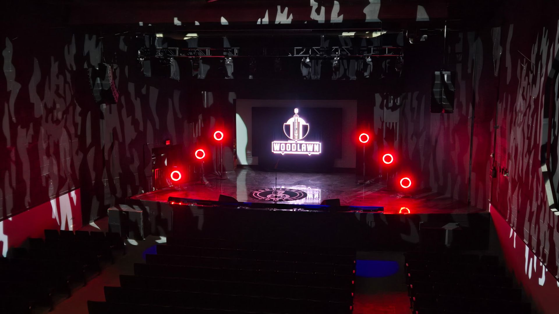 A stage illuminated by red lights and a Woodlawn Comedy logo on a screen in center.