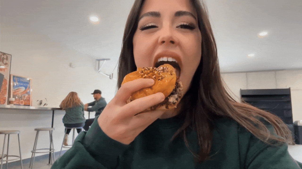 Close-up of reporter Abbey with long brown hair wearing a green top, about to bite into a sandwich with sesame seeds. Two people sit on stools at a counter in a brightly lit café.