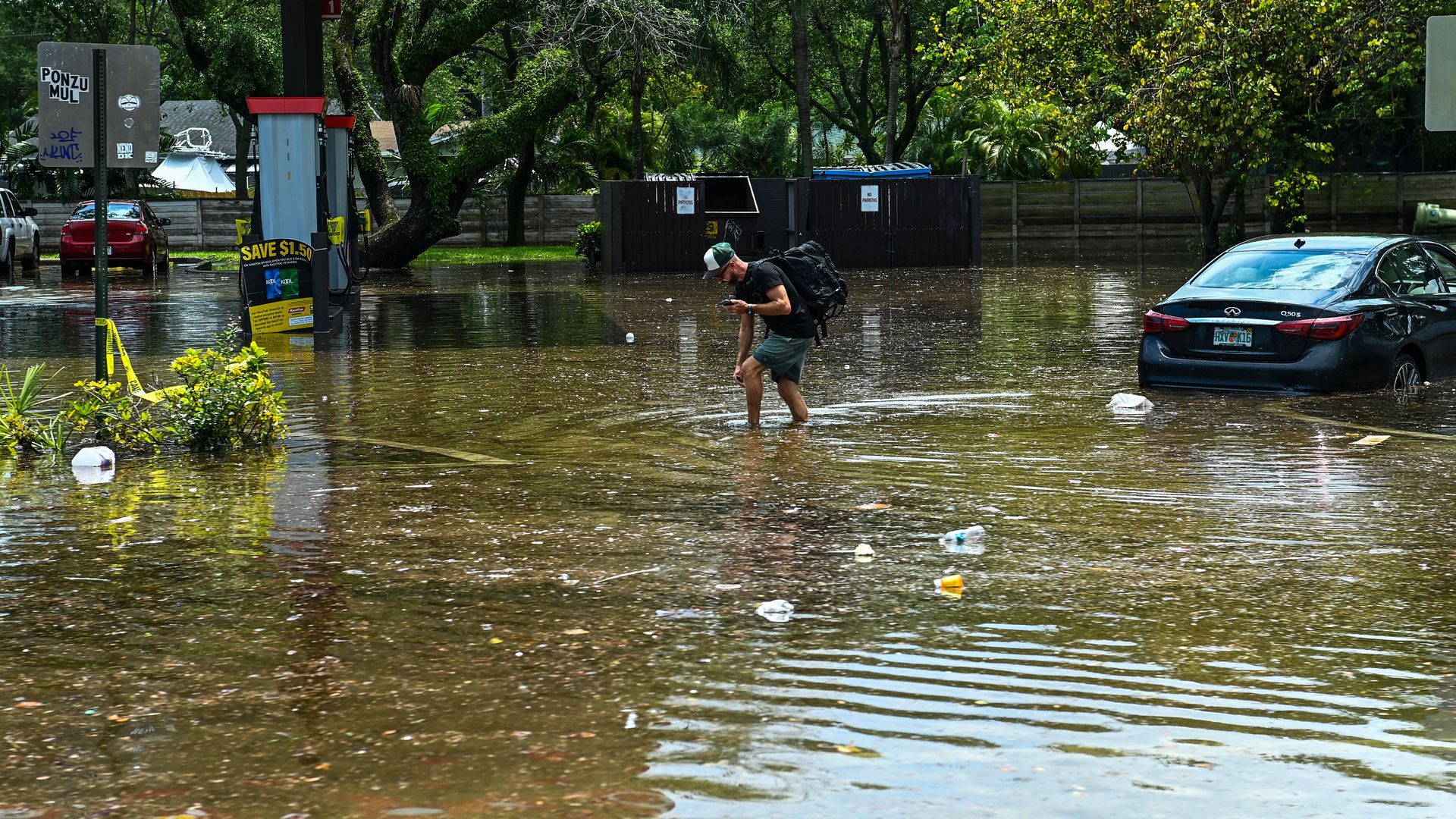 A man wades through a flooded street, near a partially submerged car and gas station
