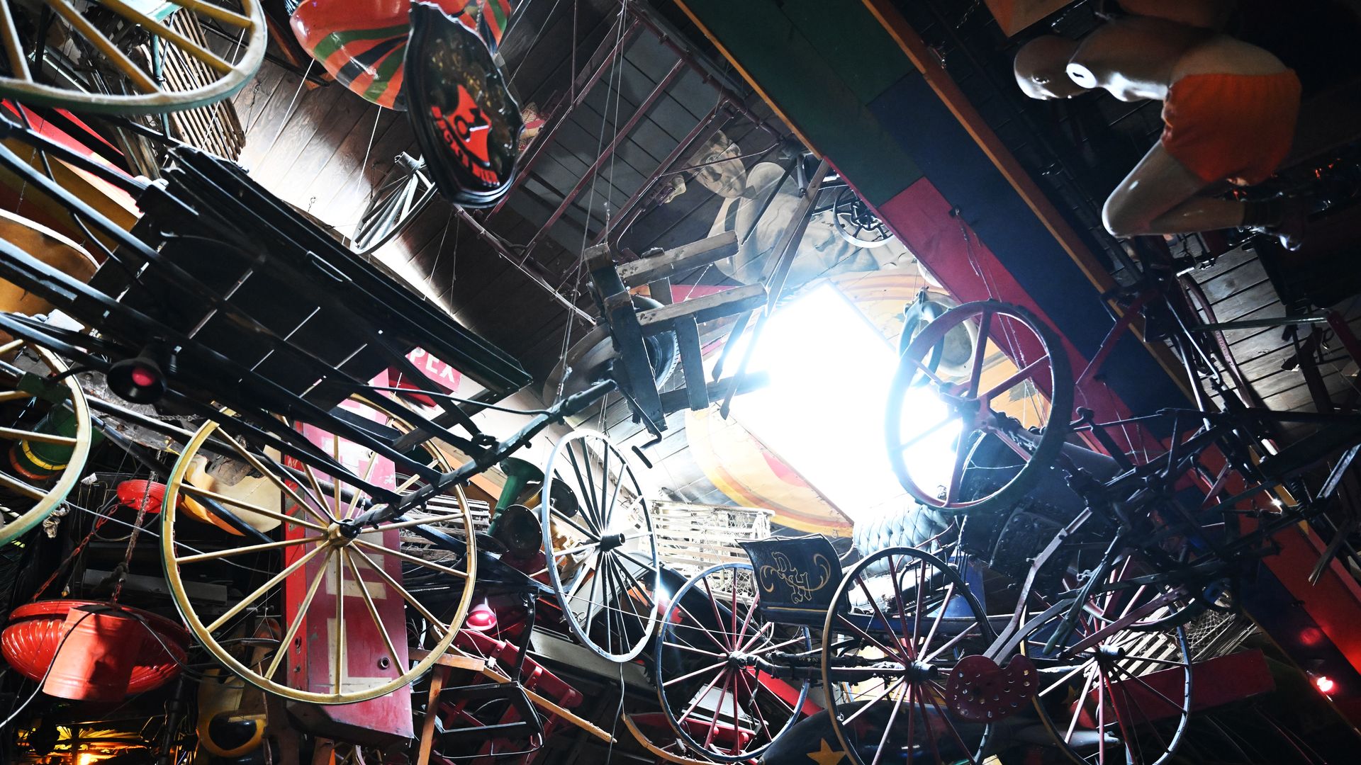 Ceiling cluttered with hanging vintage bicycles and wheels in red, blue, yellow; light pours from a skylight, creating a chaotic colorful display with a person in orange shorts on the right.