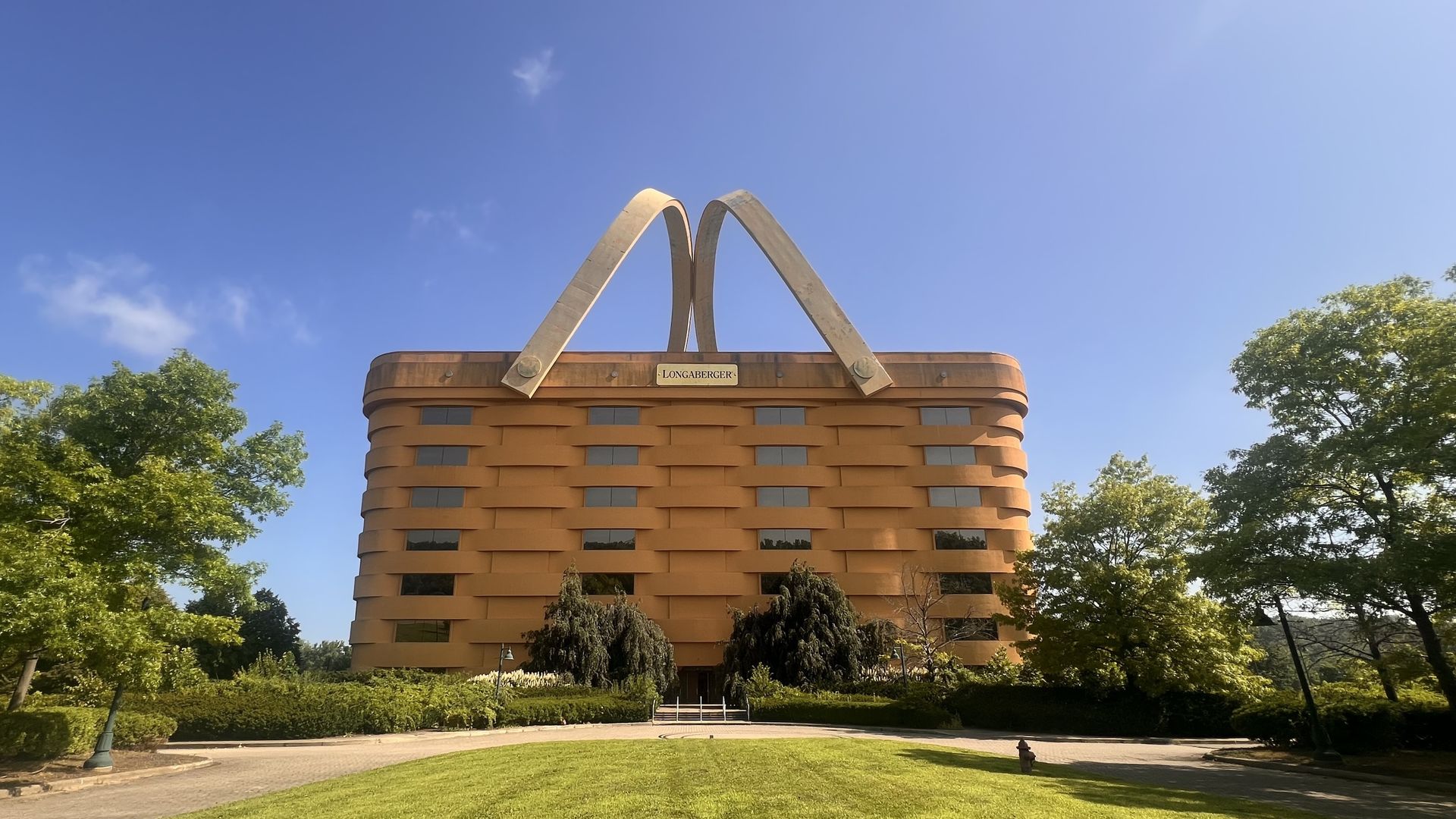 Tan brick building with two large metal arches on its roof forming an M; a plaque reads Longaberger, set against a clear blue sky with green trees in the foreground.
