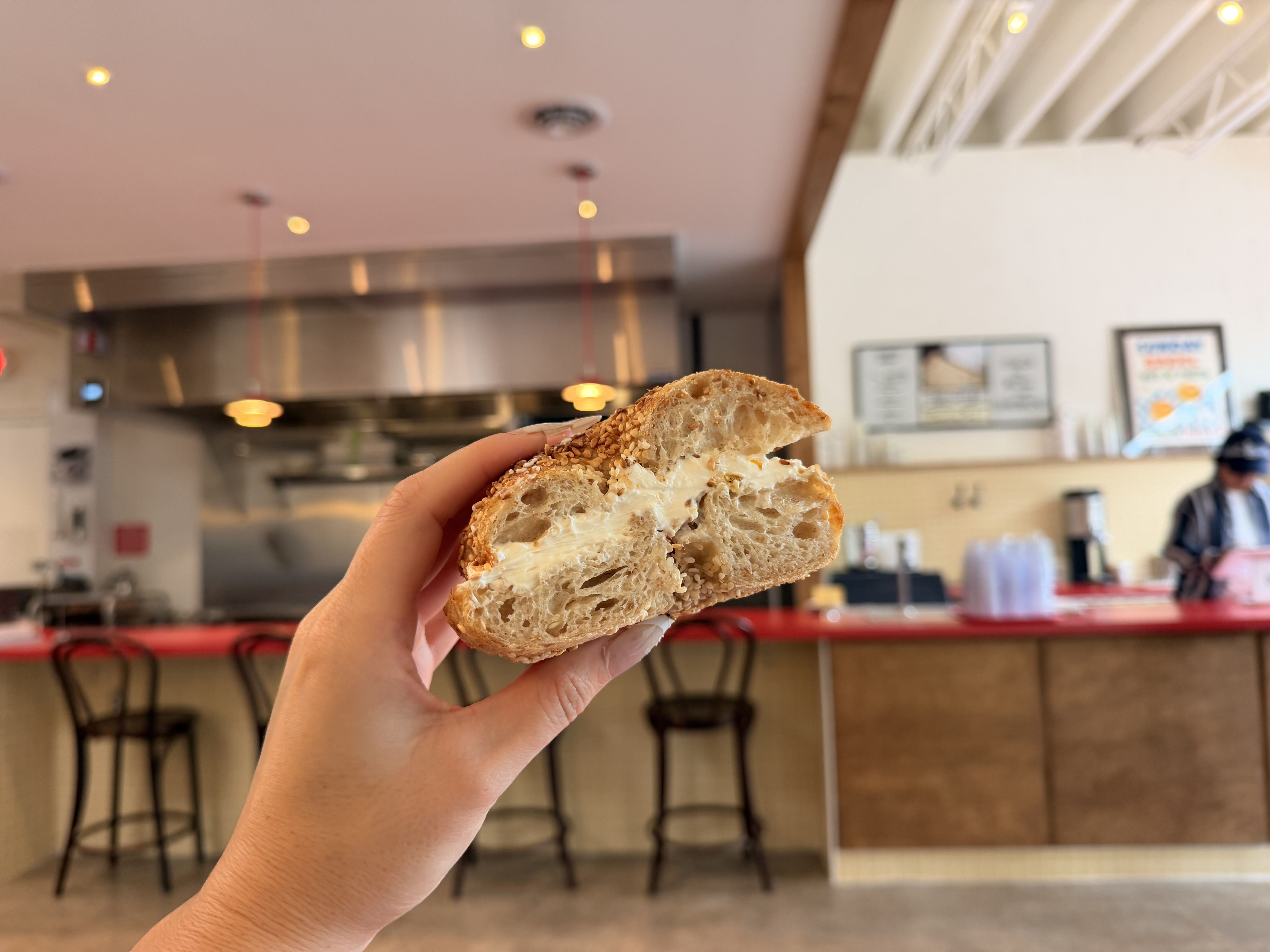 Close-up of a hand holding a half-eaten sesame seed bagel with cream cheese, in a bright cafe with a stainless steel kitchen and blurred patrons in the background.