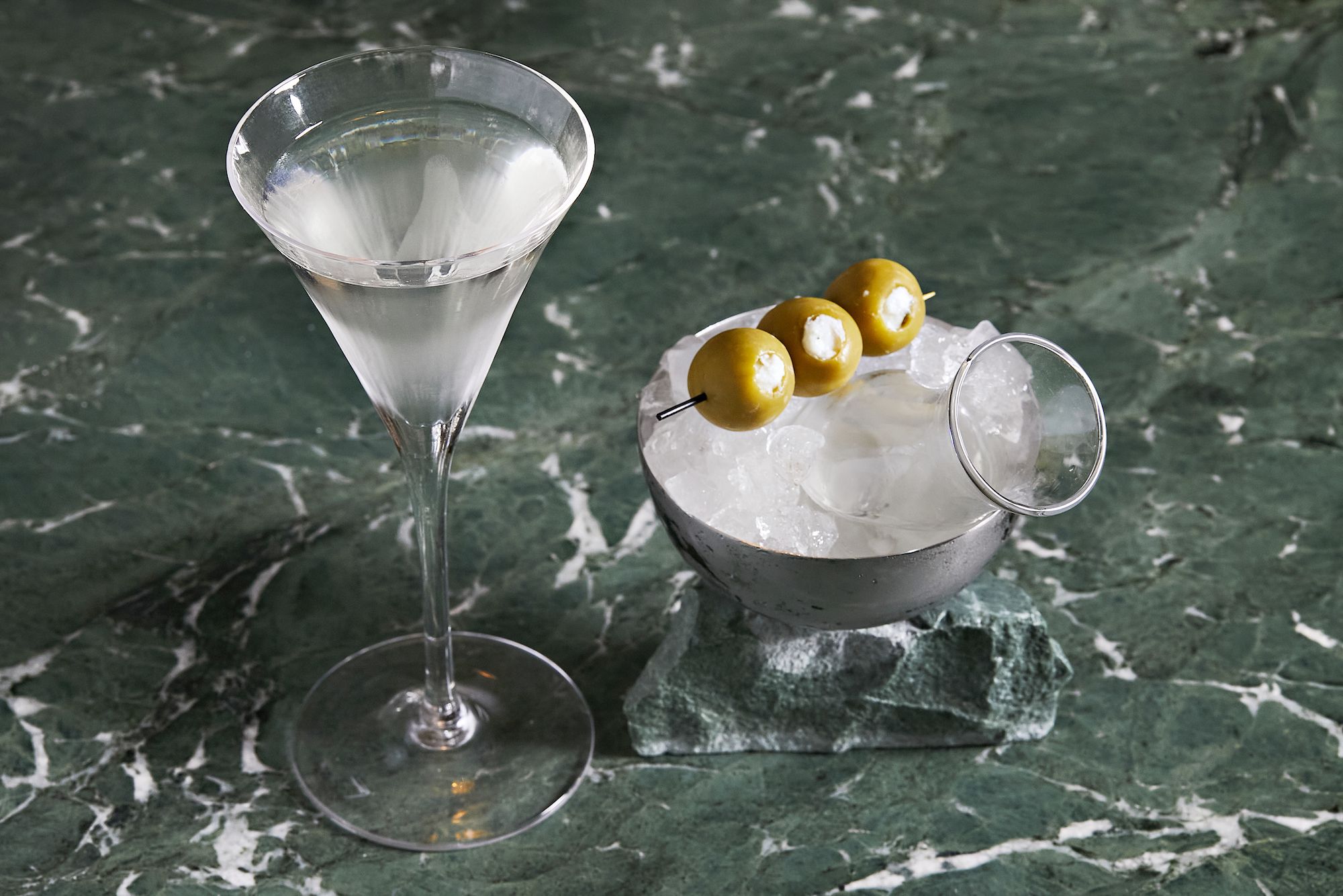 Clear martini cocktail in a glass with three stuffed green olives on a skewer resting on a bowl of ice, on a green marble surface.