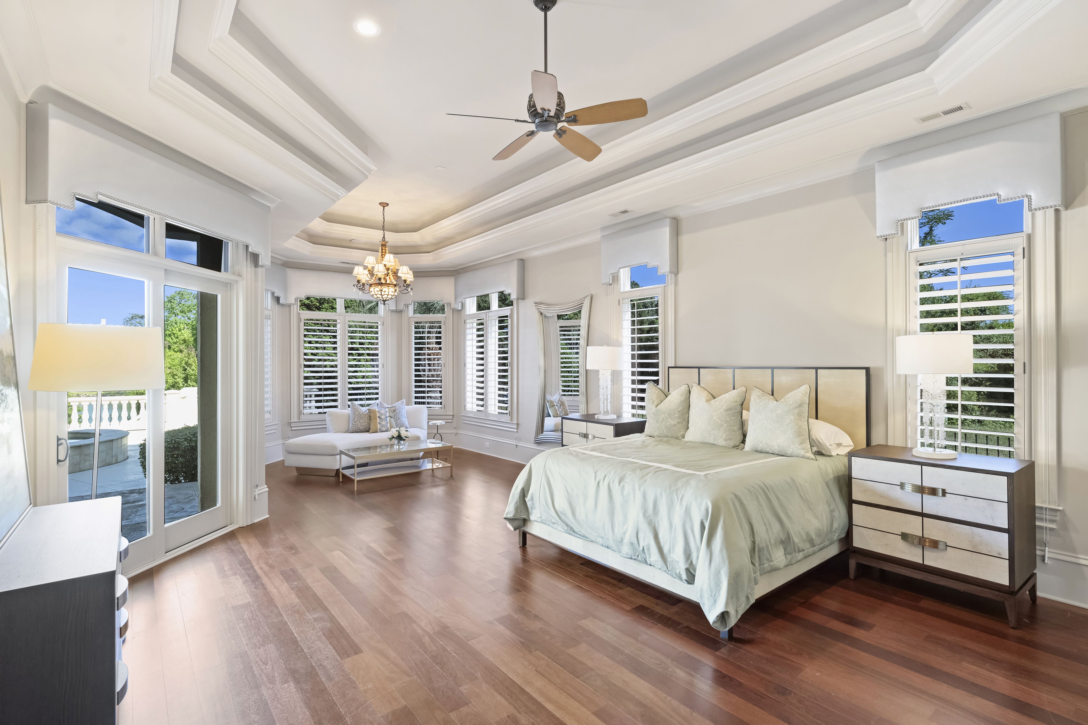 Bright bedroom with tray ceilings, white crown molding, and hardwood floors. A ceiling fan and chandelier overlook a seating area. Large windows with shutters and a balcony door.