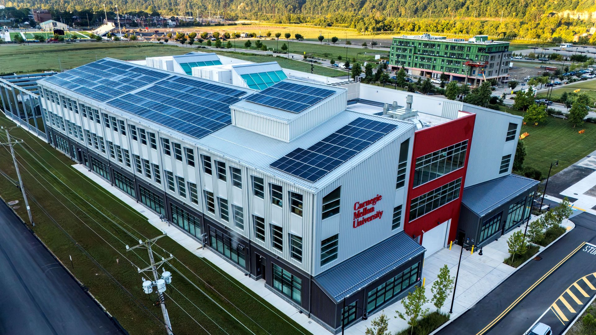 Aerial view of a modern campus building with a large solar roof, white/gray exterior, red accent wall, and Carnegie Mellon University signage; green hills and additional buildings in the distance.