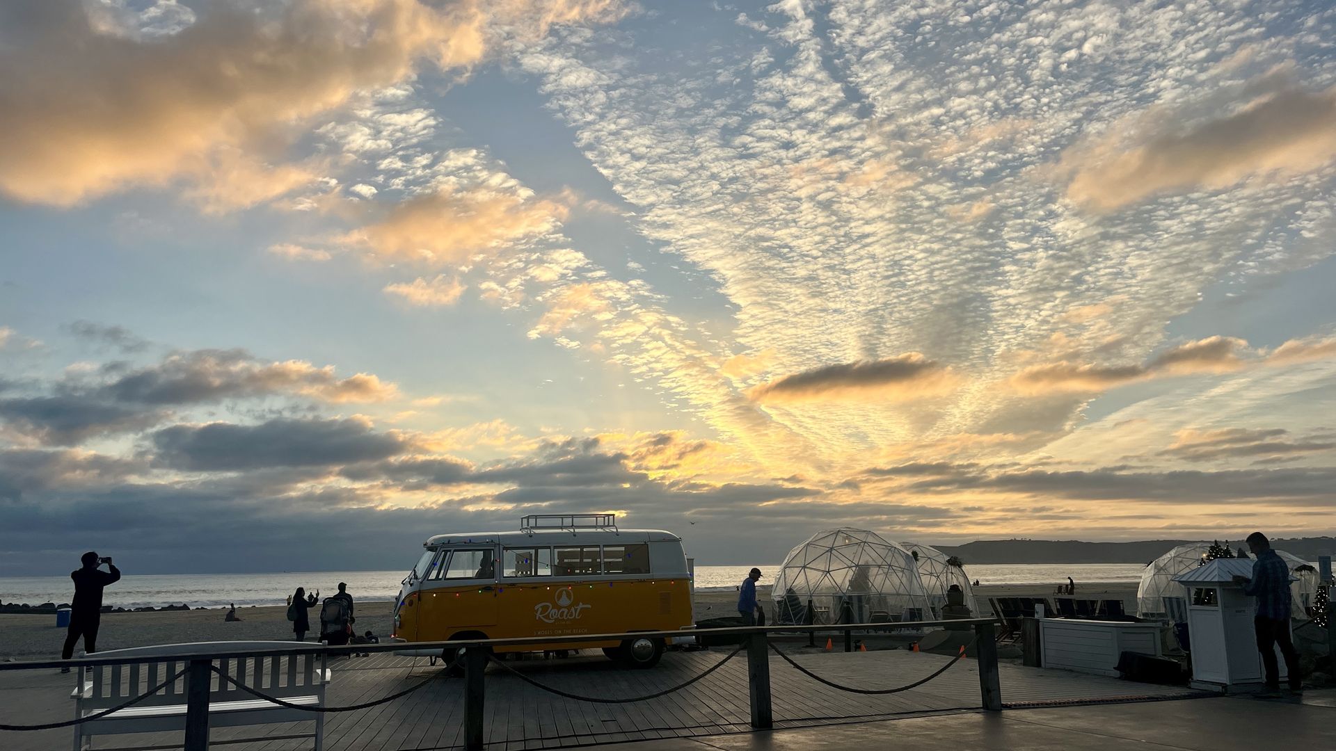 Sunset beach scene with a yellow VW van and igloo-shaped seating areas. Silhouettes of people near ocean with dramatic clouds and golden sky.