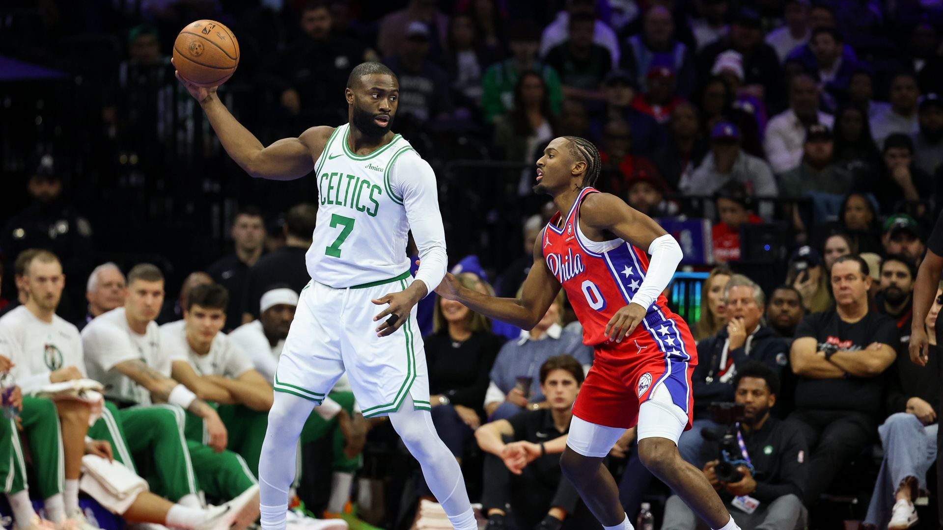 Jaylen Brown of the Celtics, wearing a white jersey with green lettering, holds a basketball in his extended right hand away from Tyrese Maxey of the 76ers, who's wearing a red-white-and-blue jersey and eyeing the ball. 