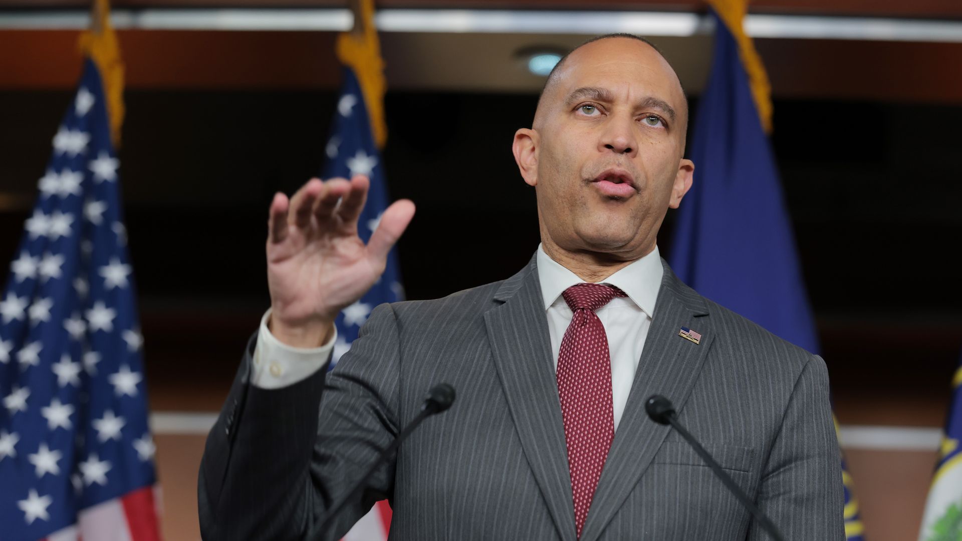 House Minority Leader Hakeem Jeffries, wearing a gray pinstripe suit and standing in front of a row of American flags in a wood-paneled room.