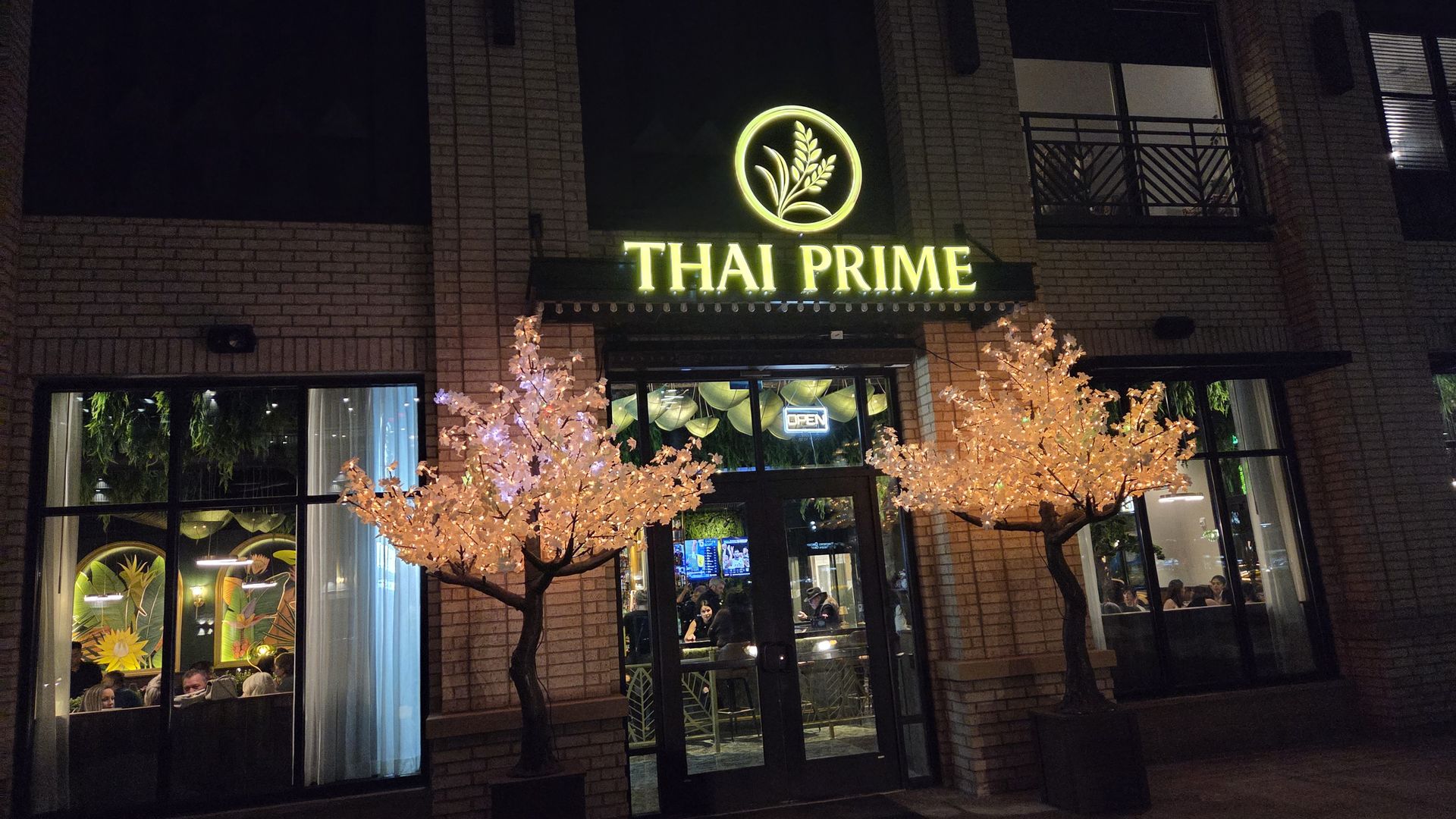 Night view of Thai Prime restaurant entrance with illuminated signage, two lit decorative trees, and large windows showing people dining inside.