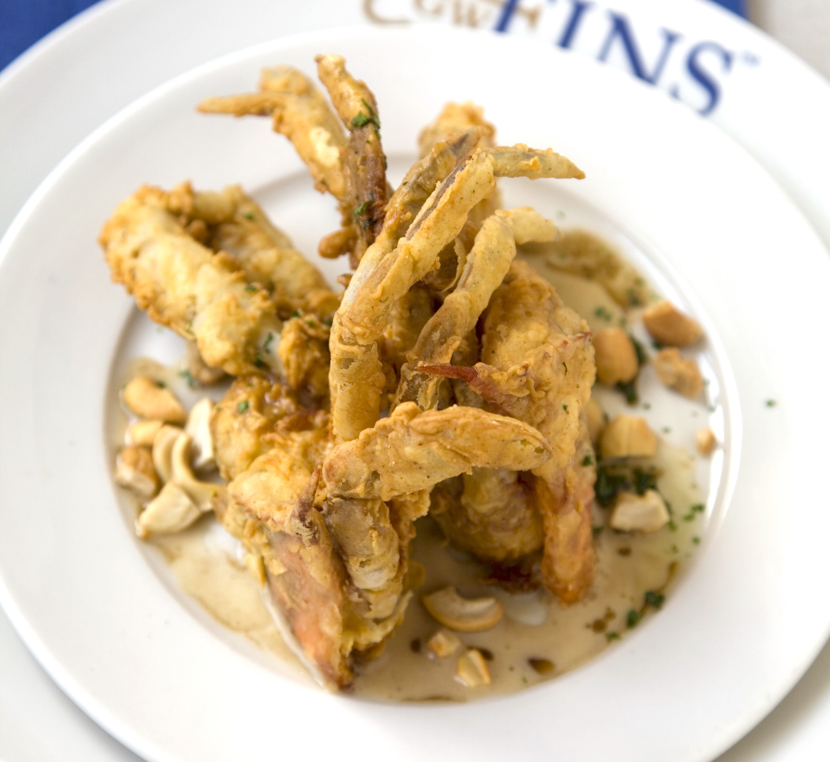 Photo shows a deep-fried soft shell crab on a white plate with sauce under it.