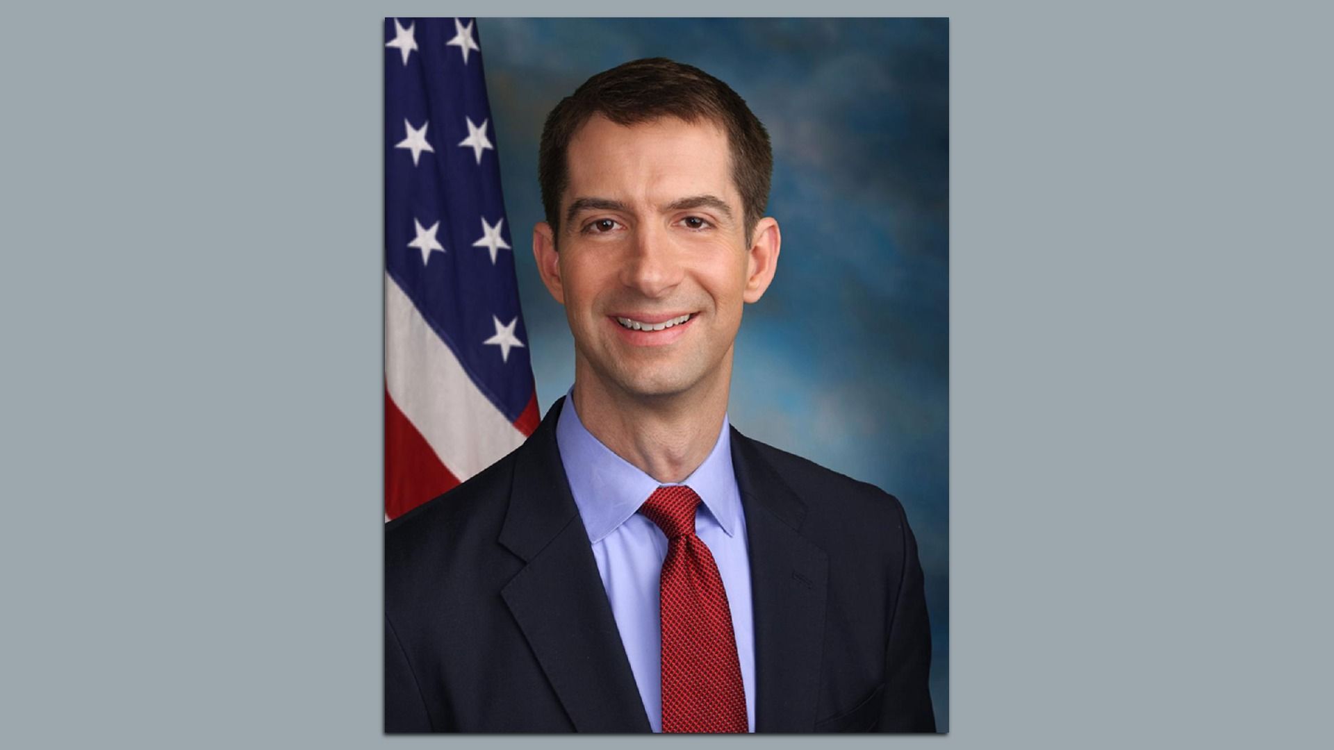 Portrait of a smiling man in a dark suit with a light blue shirt and red tie, standing in front of an American flag against a blue gradient background.