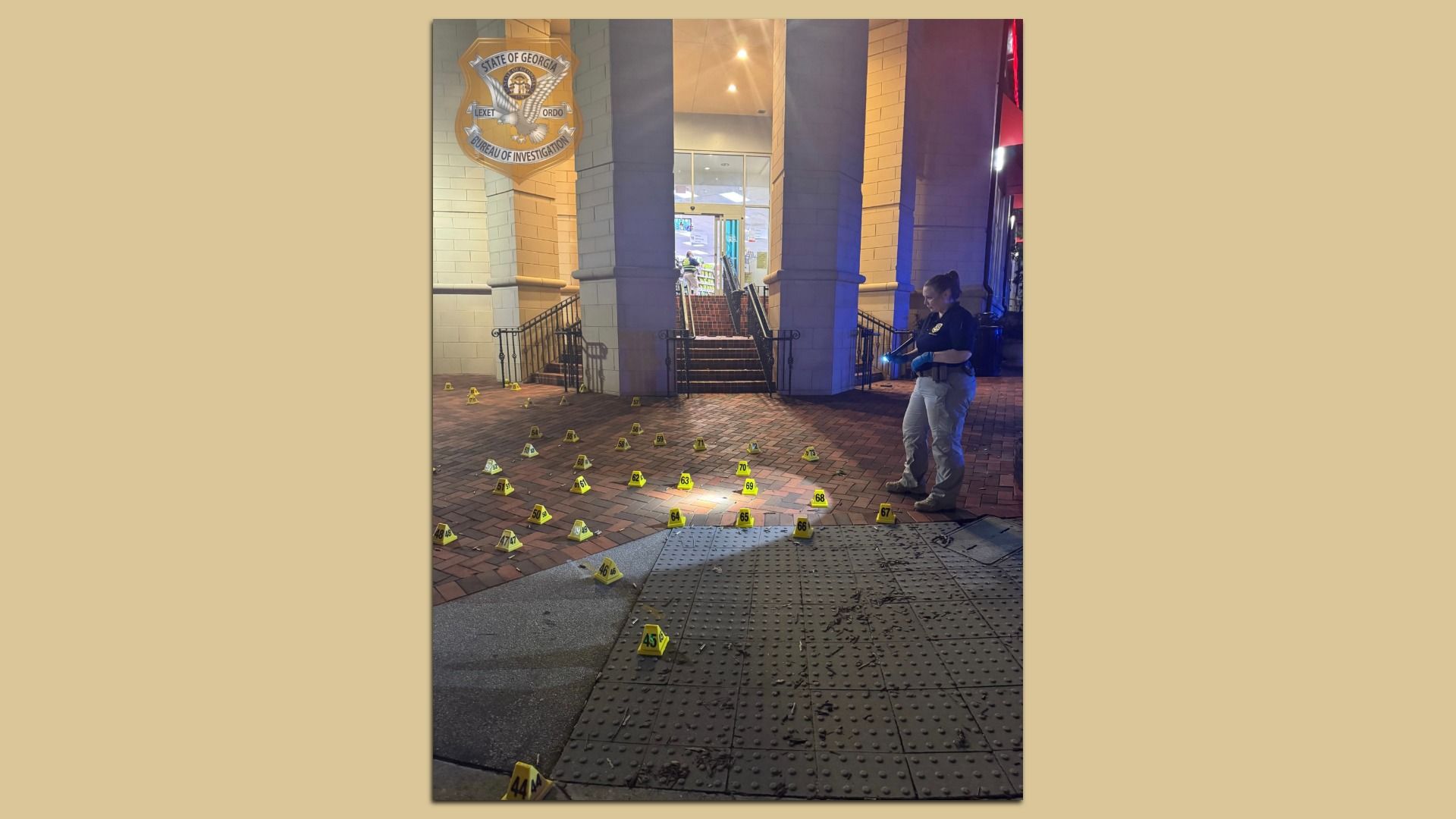 Investigator with gloves and flashlight examines numerous numbered yellow evidence markers scattered on brick pavement near building entrance at night.