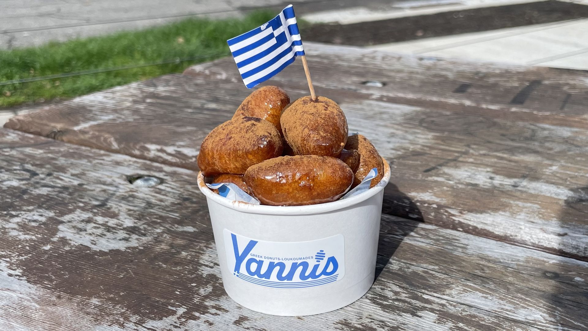 White cup labeled "Yannis" filled with golden brown loukoumades dusted with cinnamon sugar; a small Greek flag on a toothpick stands among the stacked donuts, on a weathered wooden table outdoors.