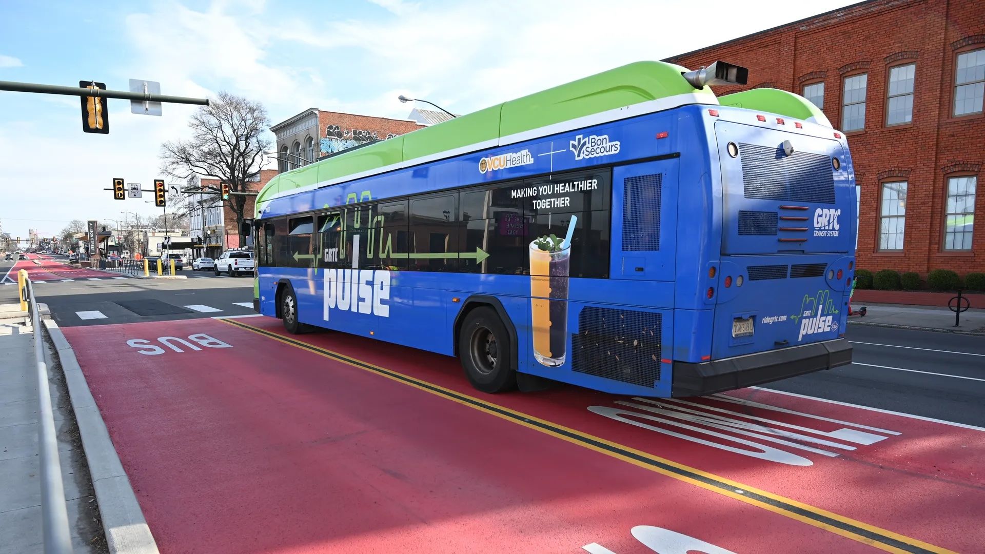 Blue and lime-green GRTC Pulse bus in a red bus lane on a city street; the side shows a large Pulse logo and a beverage ad. Brick buildings, traffic lights, and a crosswalk in the background.