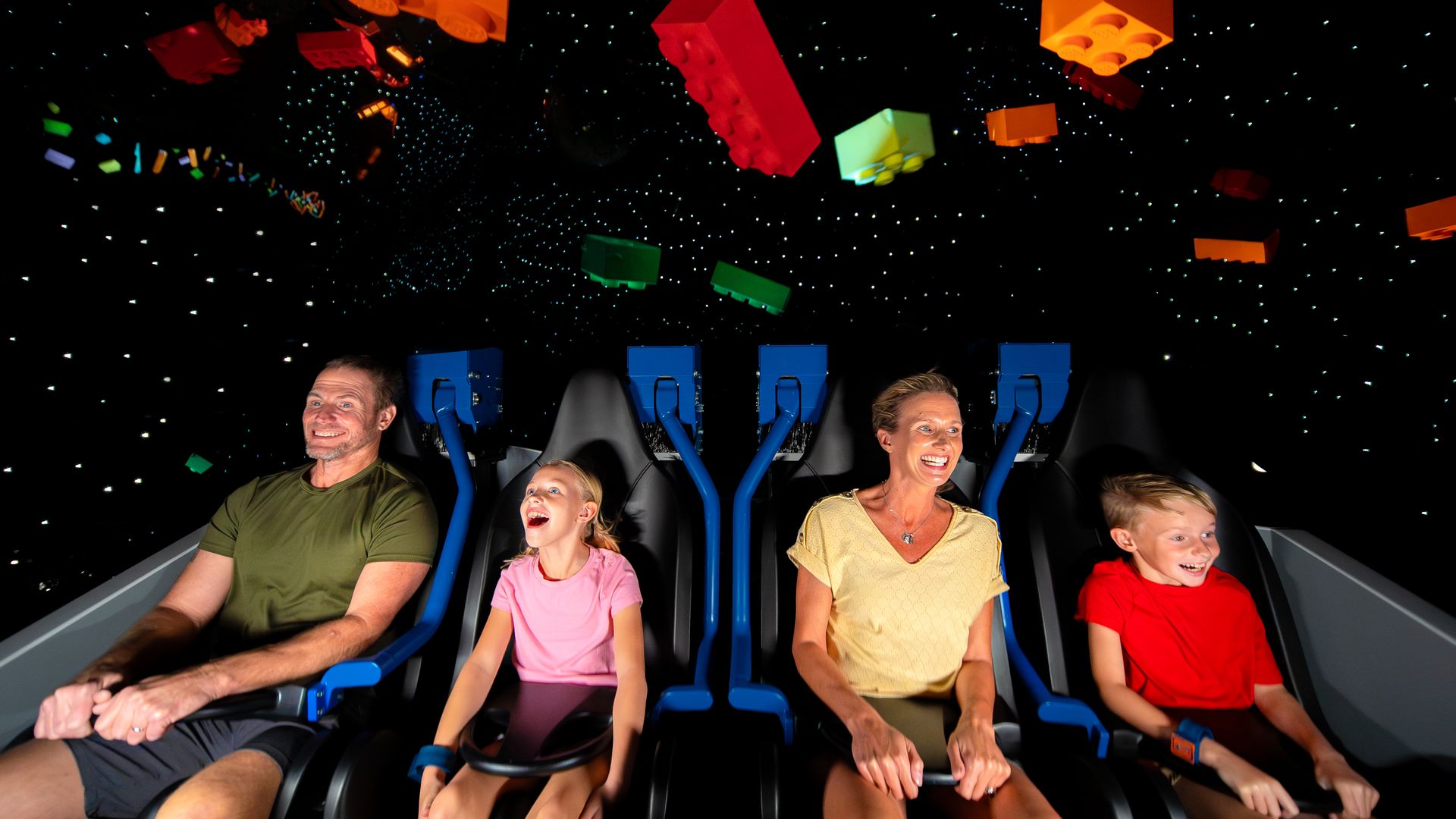 A family of four smiling and seated in a dark amusement ride with large colorful Lego blocks suspended above them.