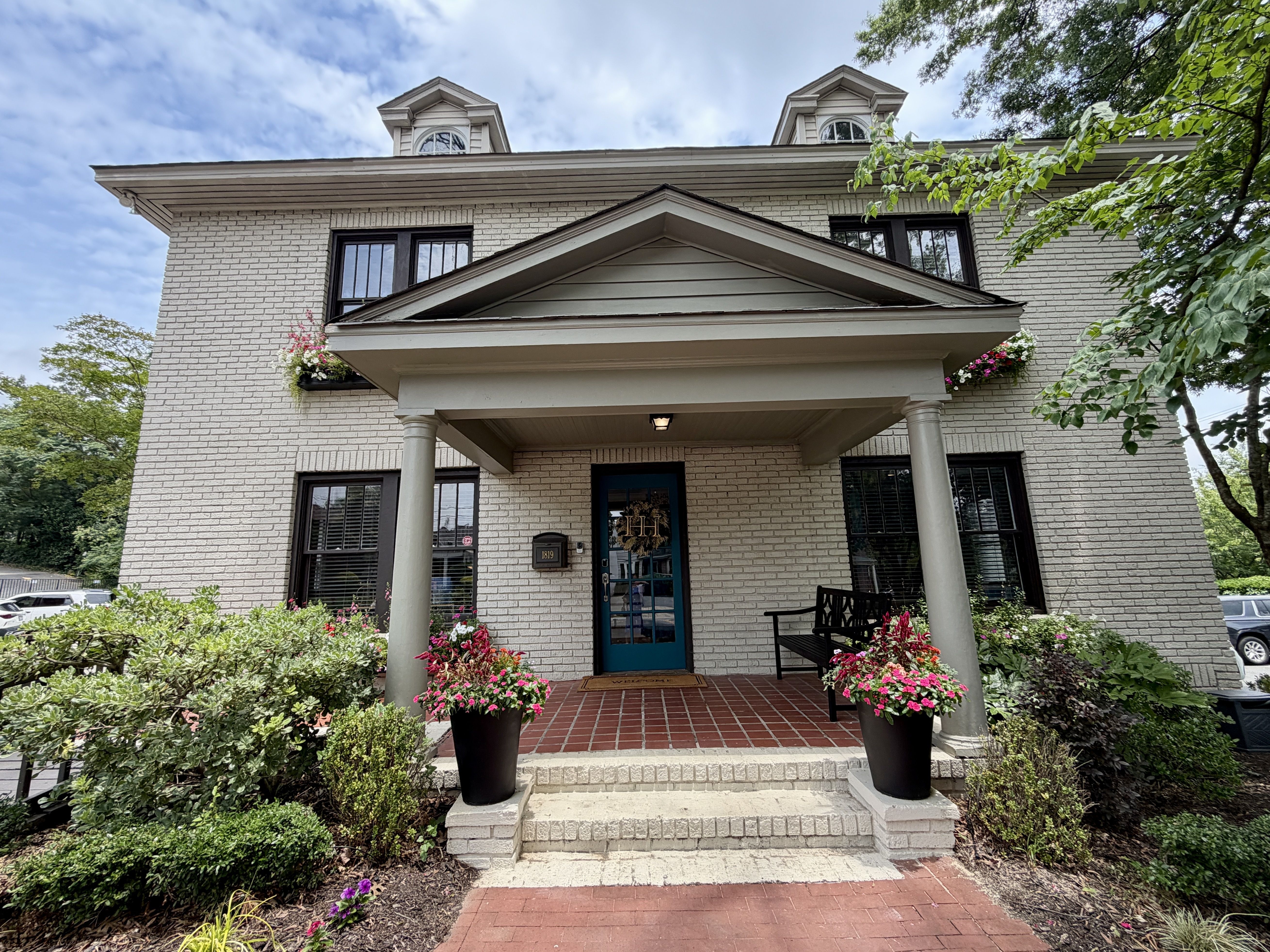 Two-story light-brick house with a centered porch and triangular pediment, flanked by two dormer windows. A blue front door, dark-framed windows, and pink flowering planters with greenery.