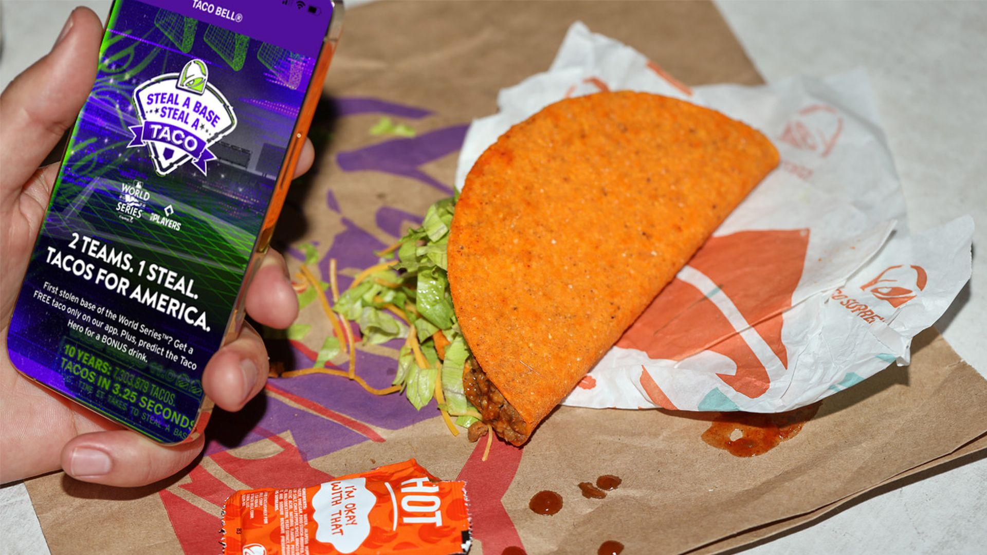 Hand holding phone with Taco Bell promotion on it and a Taco Bell taco, sauce packet