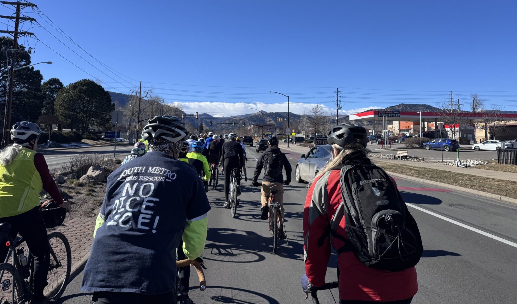 Group of cyclists riding on a city street under clear blue sky, with mountains in the background and a red and white gas station visible on the right side.