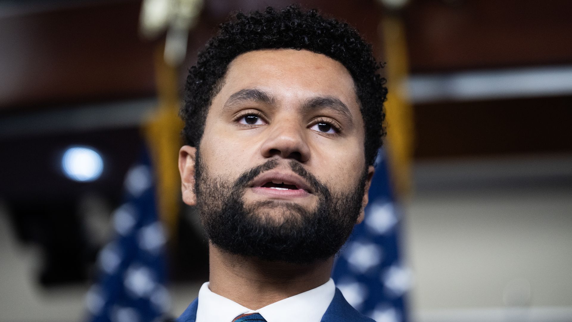 Close-up of a man with curly hair and beard wearing a blue suit, white shirt, and green plaid tie, speaking at a podium with blurred American flags in the background.