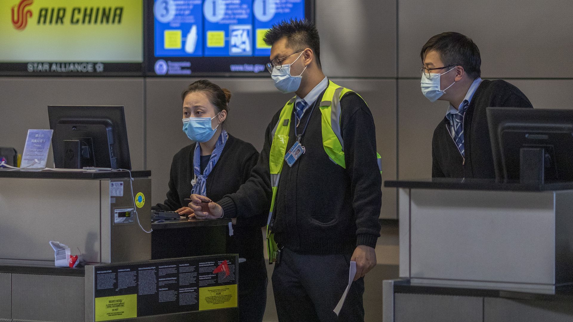 In this image, three airport employees stand at a baggage claim while wearing face masks.