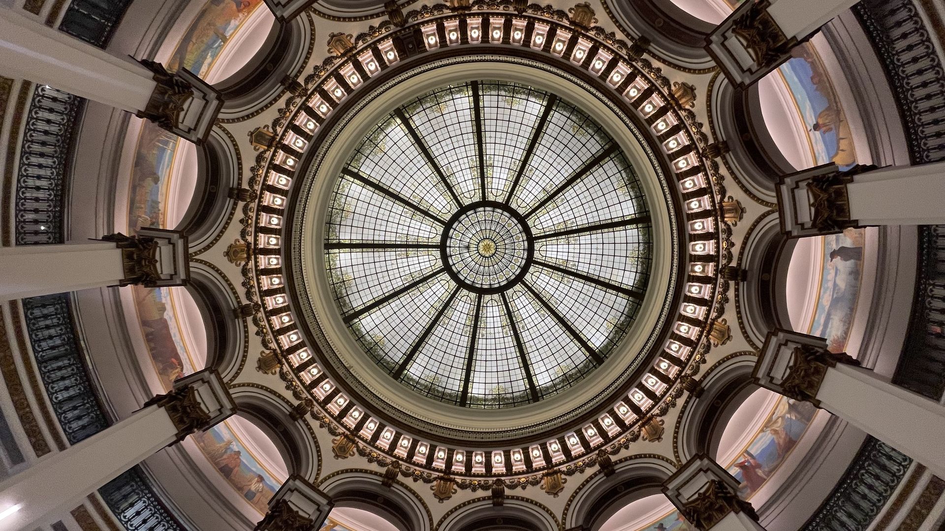 Interior view of a grand domed ceiling with a circular stained-glass skylight. Black metal radial spokes form a sunburst, surrounded by gold-accented arches, statues, and mural-adorned balconies.
