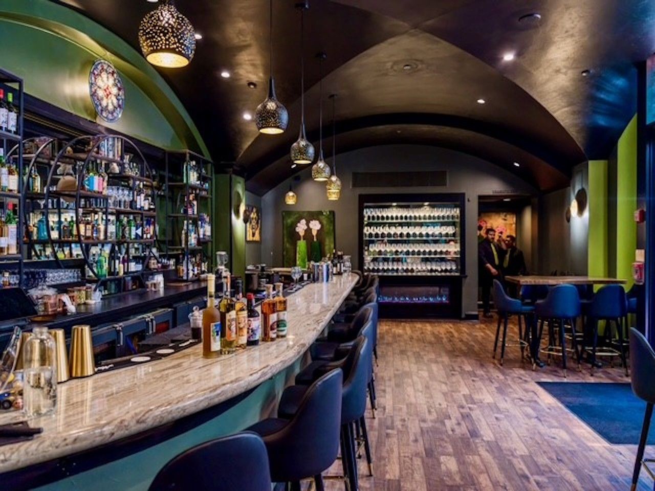 Stylish bar interior with a long marble counter, bottle displays, hanging pendant lights, arched ceiling, blue chairs, and a back refrigeration unit; warm, modern ambiance.