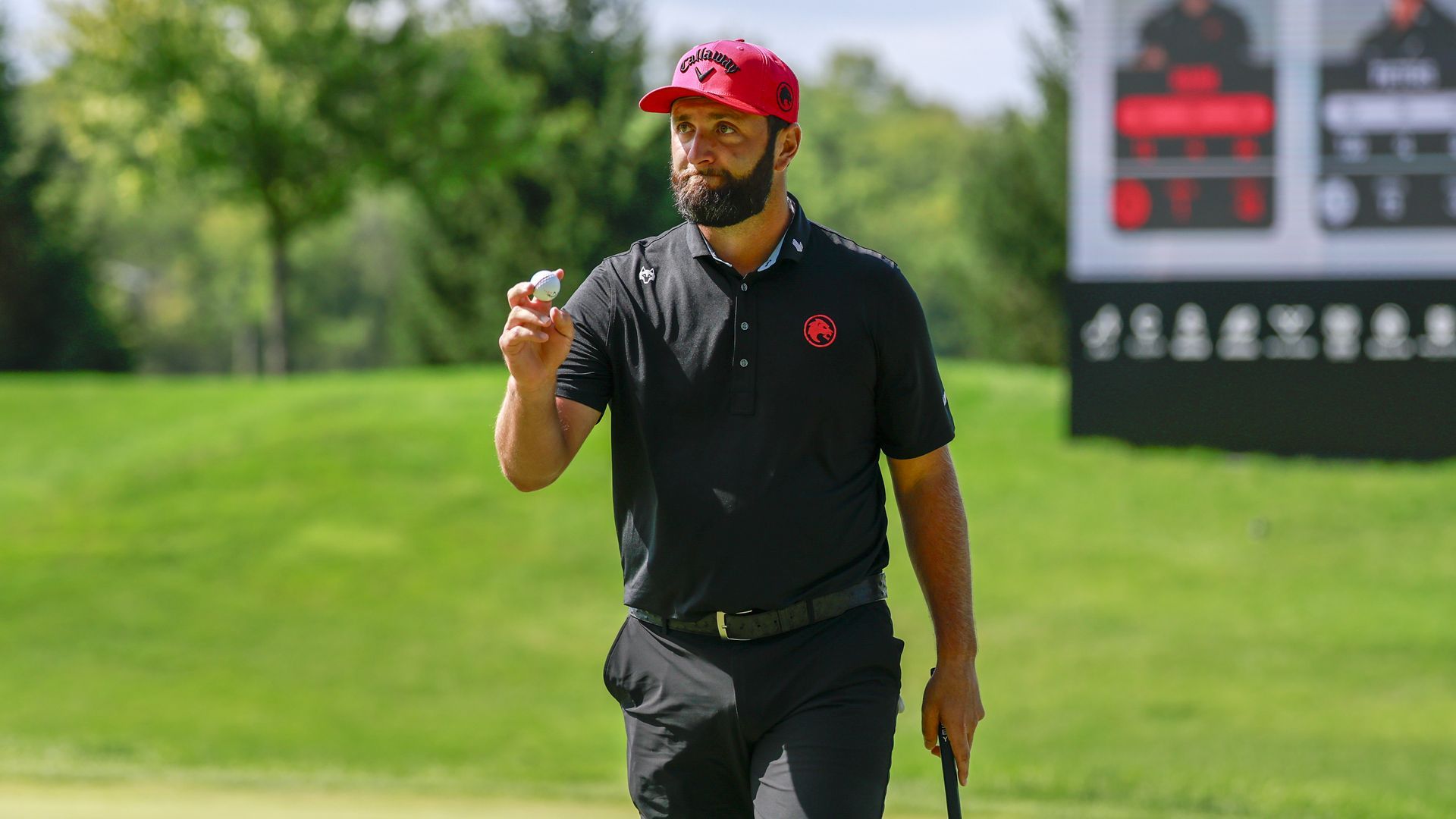 Bearded man in a red cap and black polo on a sunny golf course, holding a golf ball in his right hand as a scoreboard blurs in the background.