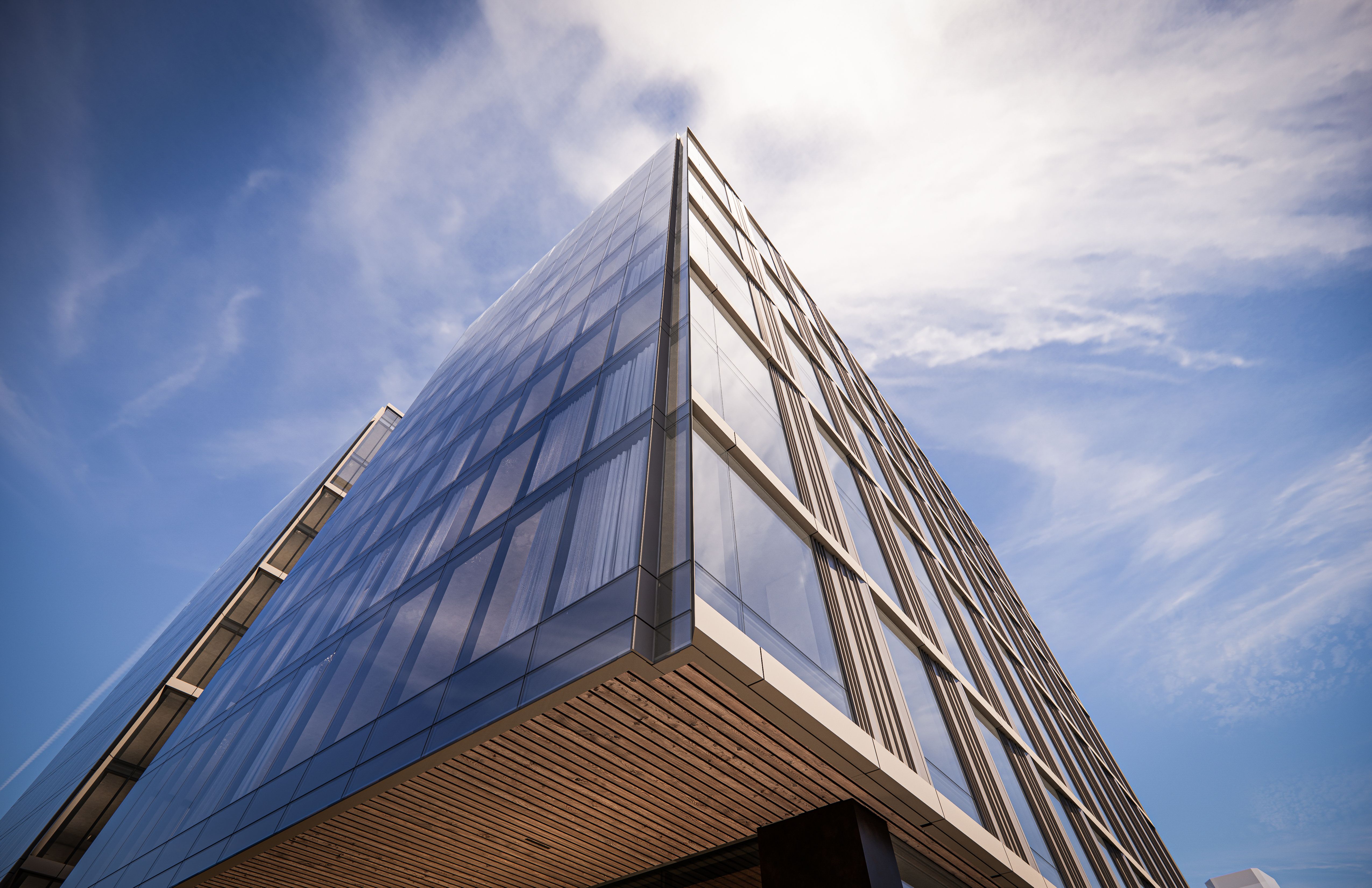 Low-angle view of a tall glass office building with reflective blue windows, metal frames, and a wooden-louvered overhang, set against a bright blue sky with scattered white clouds.