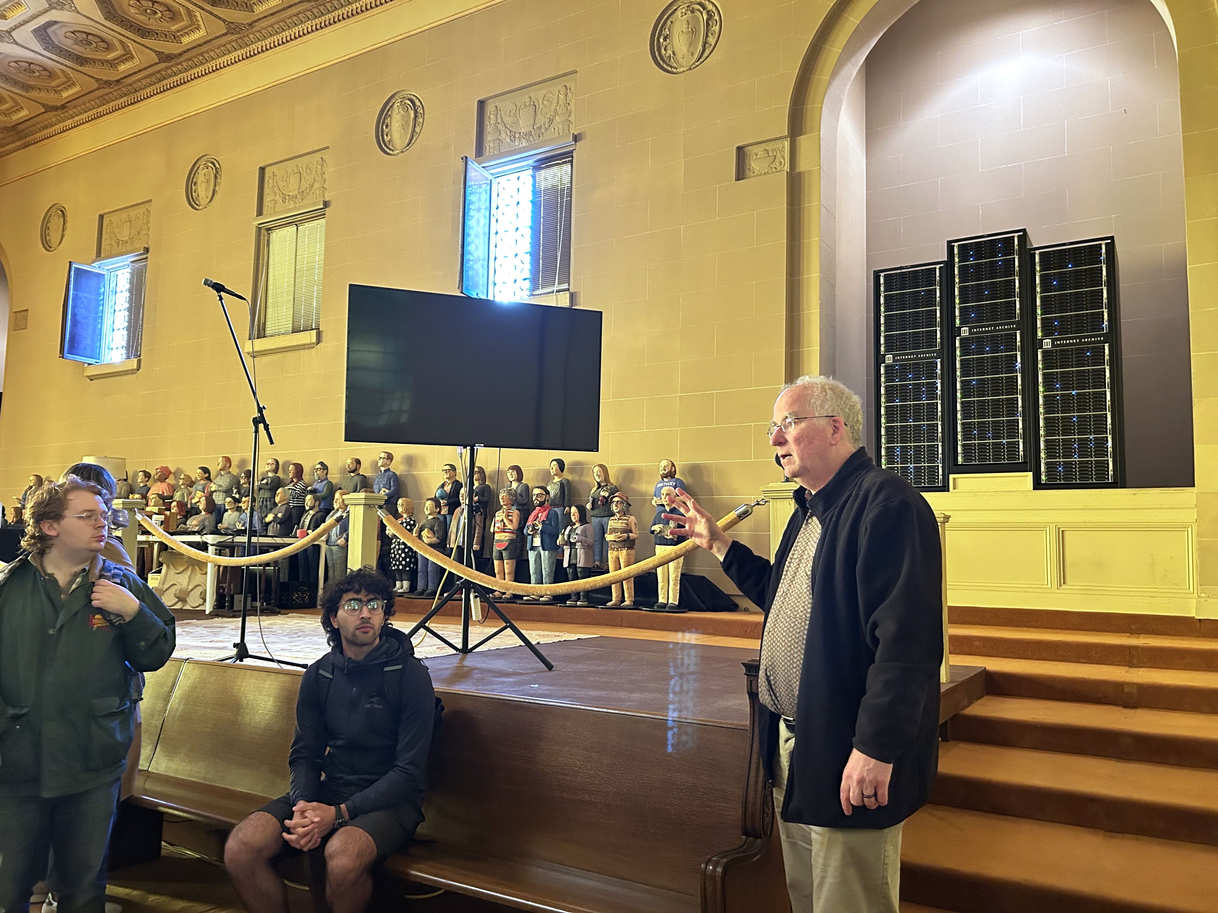 Man in black jacket speaking inside a hall with yellow walls, wooden bench, and large server racks. Sculpture figures and a black monitor on a stand are in the background.
