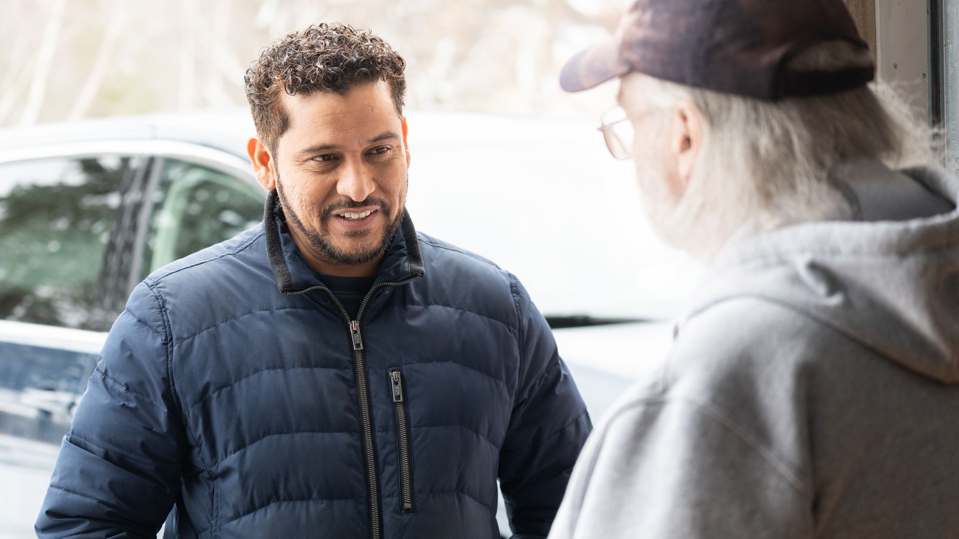 Two men chat outdoors beside a car: left man in a navy puffer jacket with short curly hair, right man in a gray hoodie and cap.