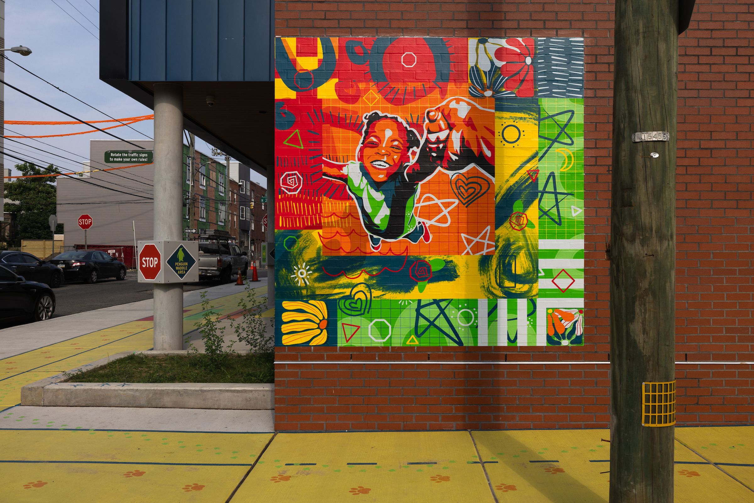 Colorful mural on a brick wall depicting a smiling child reaching out, surrounded by bright red, orange, green, and yellow geometric and floral patterns, with a wooden pole nearby.