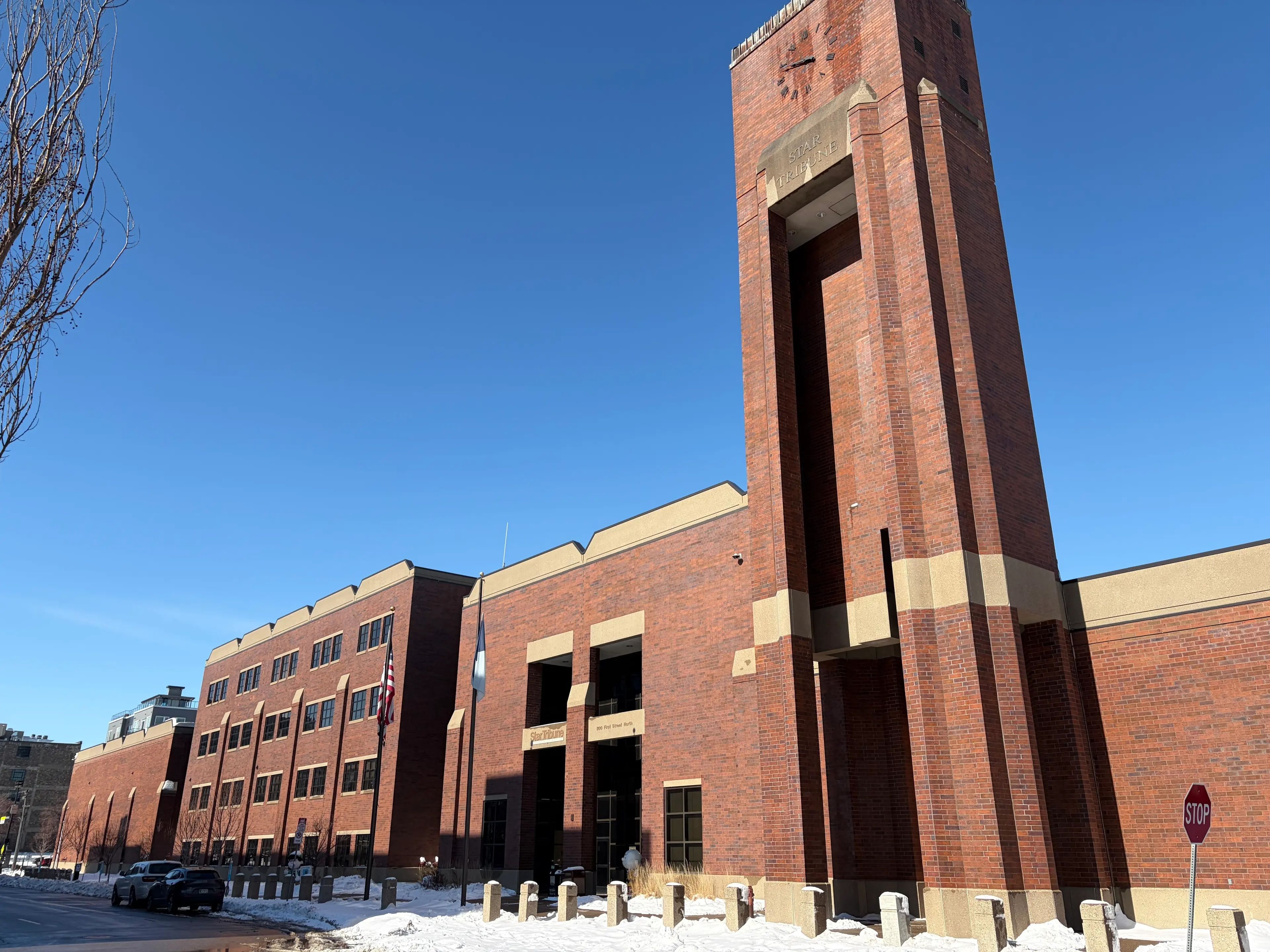 The Star Tribune building, a red-brick complex with a tall square clock tower, under a clear blue sky; snow on the ground, flags at the entrance, and a stop sign by the street.