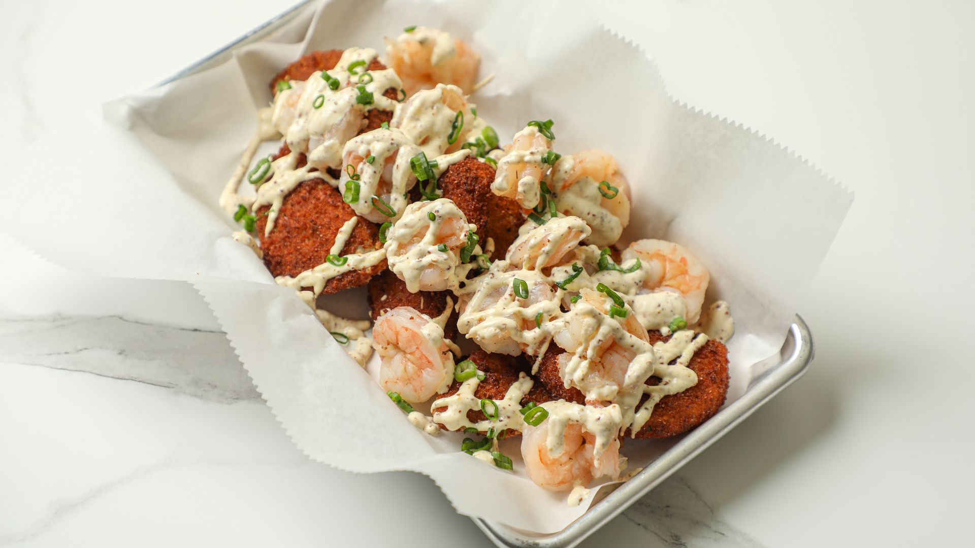 A metal tray lined with parchment paper holds fried croquettes topped with shrimp, drizzled with a creamy white sauce, sprinkled with chopped green onions, on a marble surface.