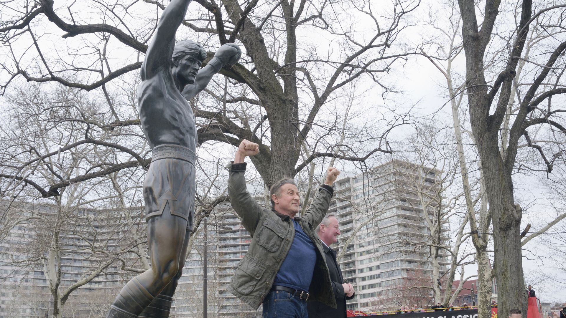 Sylvester Stallone standing with his arms and fists in the air like a winning boxer, in front of the Rocky Statue in Philly, which has the same pose. 
