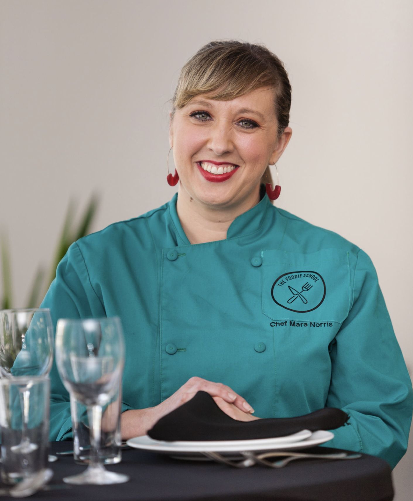 Smiling woman in a teal chef jacket with "Chef Mara Norris" embroidered, sitting at a table set with plates, a black napkin, and empty glasses, light background.