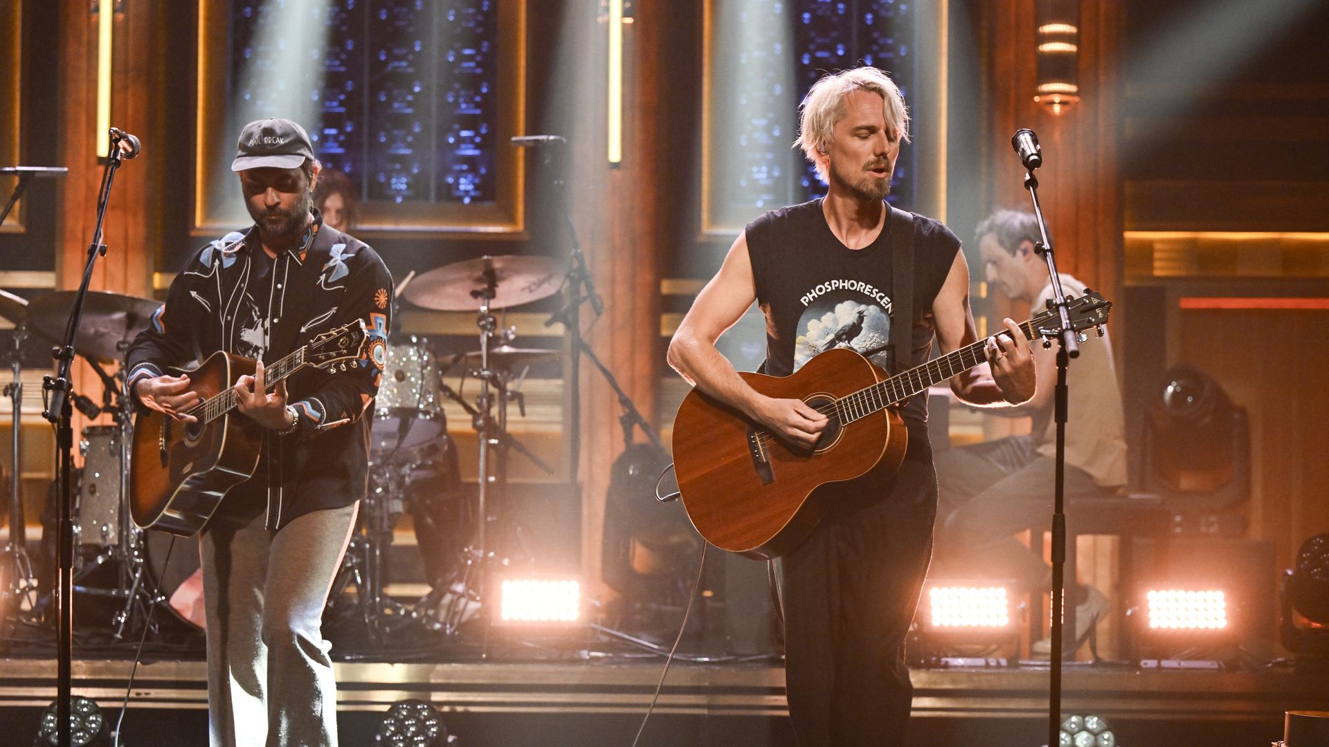 Two male musicians playing acoustic guitars on stage, one wearing a black cap and patterned jacket, the other a sleeveless black Phosphorescent shirt, warm stage lighting behind them.