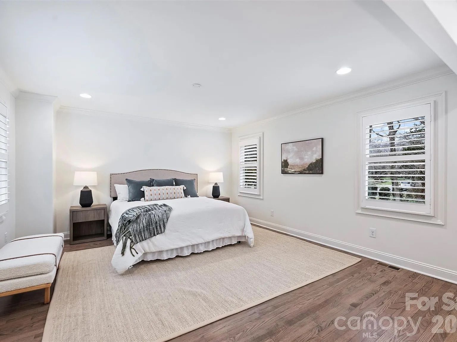 Bright, minimalist bedroom with white walls, large bed with white bedding and gray pillows, gray throw blanket, two black bedside lamps on wooden nightstands, beige rug, and two windows with white shutters.