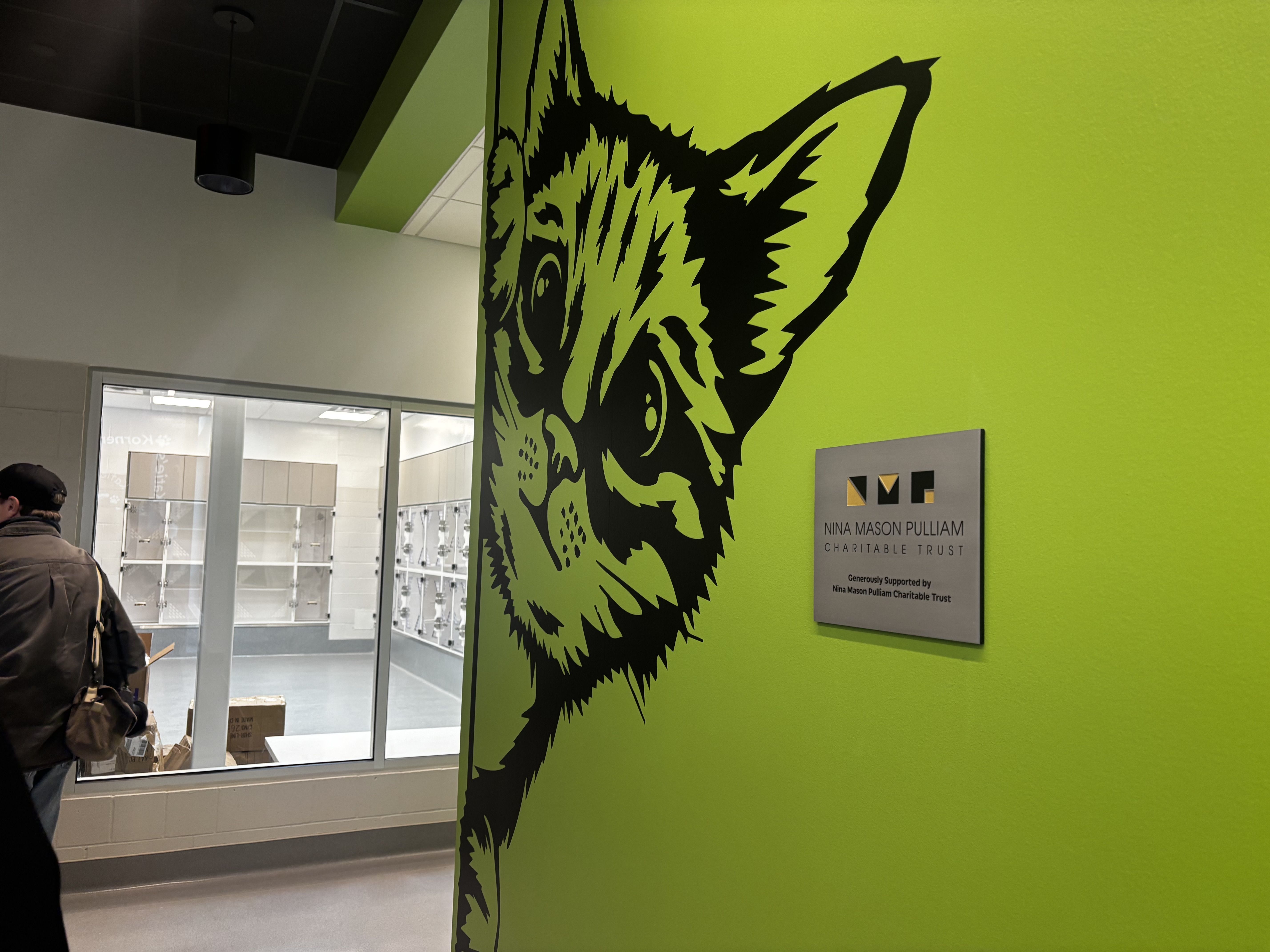 Bright lime wall with a large black animal-head graphic; a silver plaque beside it reads "Nina Mason Pulliam Charitable Trust." Glass doors and a few people are visible in a lobby.